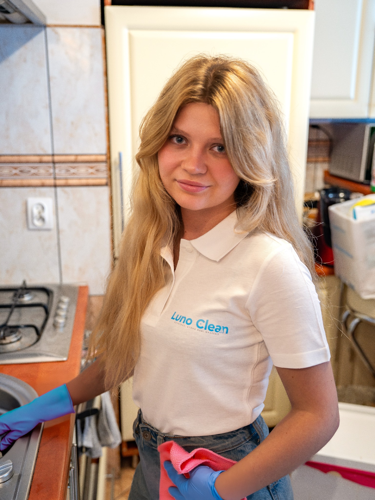 Uśmiechnięta blondynka w niebieskich rękawicach czyści kuchenkę. Na koszulce logo firmy sprzątającej 'Luno Clean'.