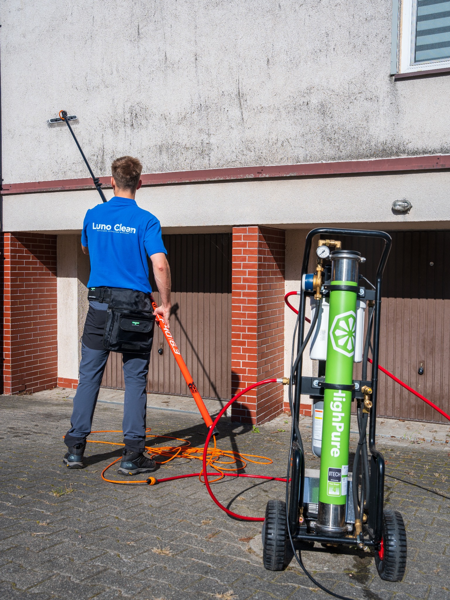 Pracownik w trakcie mycia elewacji domu za pomocą szczotki na wysięgniku i specjalistycznego urządzenia czyszczącego HighPure. Widoczny fragment ściany przed i po czyszczeniu.