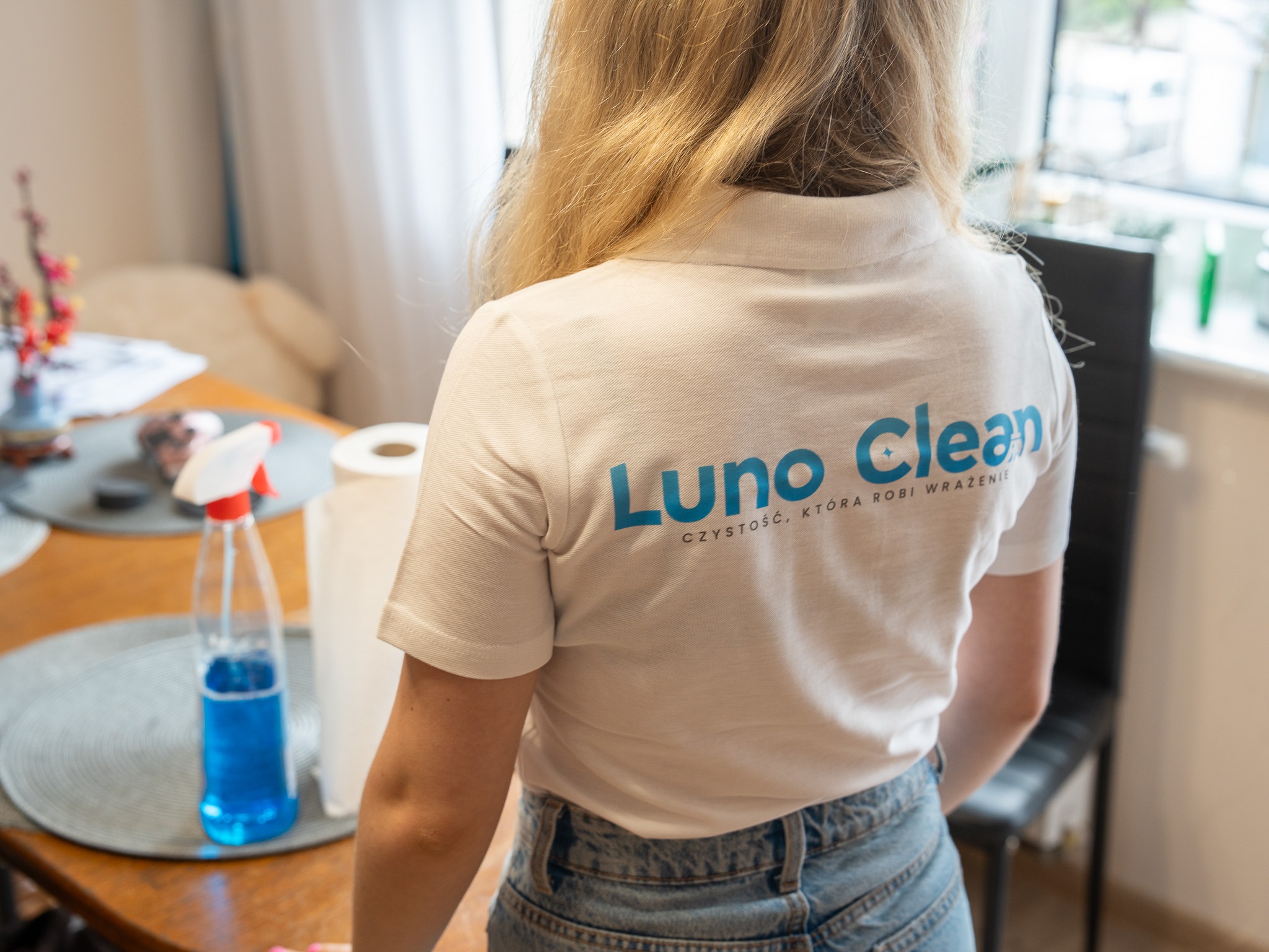 Pracownica firmy sprzątającej Luno Clean w białej koszulce polo z logo, w trakcie pracy w mieszkaniu. Na stole widoczny środek czyszczący i ręcznik papierowy.