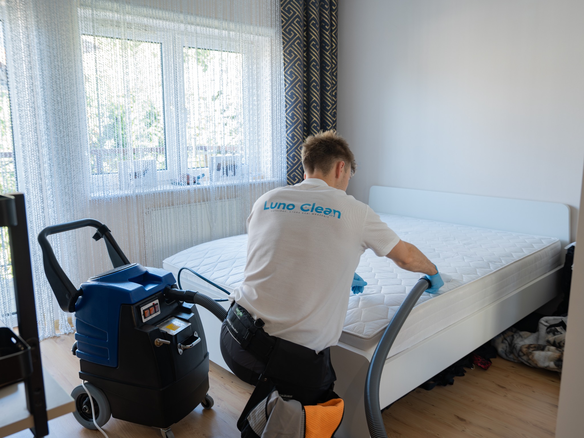 Pracownik Luno Clean czyści materac na łóżku za pomocą profesjonalnego odkurzacza piorącego w jasnym pokoju z oknem i zasłonami.