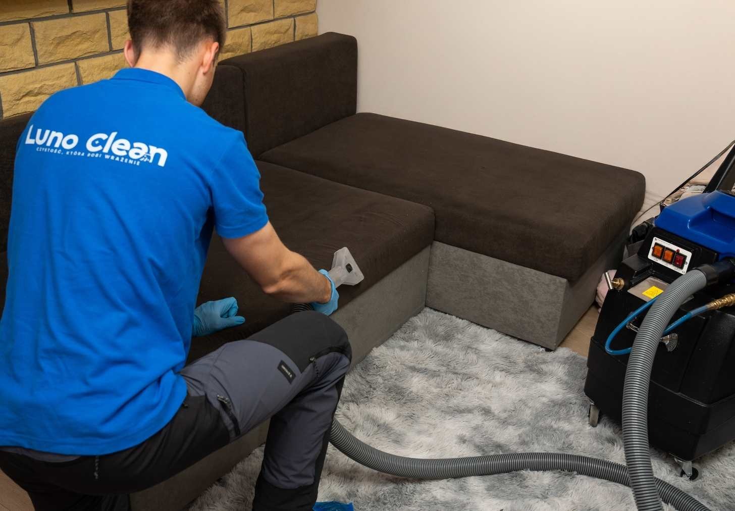 Pracownik w niebieskiej koszulce z logo czyści narożnikową kanapę parownicą. Obok stoi profesjonalny sprzęt do czyszczenia tapicerki. Dywan z długim włosiem.