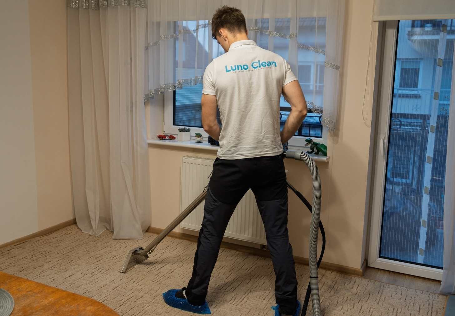 Pracownik firmy Luno Clean w trakcie prania dywanu w pokoju. Widoczne narzędzia i specjalistyczne obuwie ochronne. Jasne wnętrze z oknem i zasłonami.
