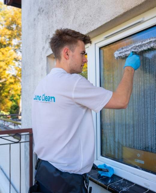 Mężczyzna w niebieskich rękawicach myje okno z zewnątrz za pomocą myjki, widoczne krople wody na szybie i logo firmy na koszulce.