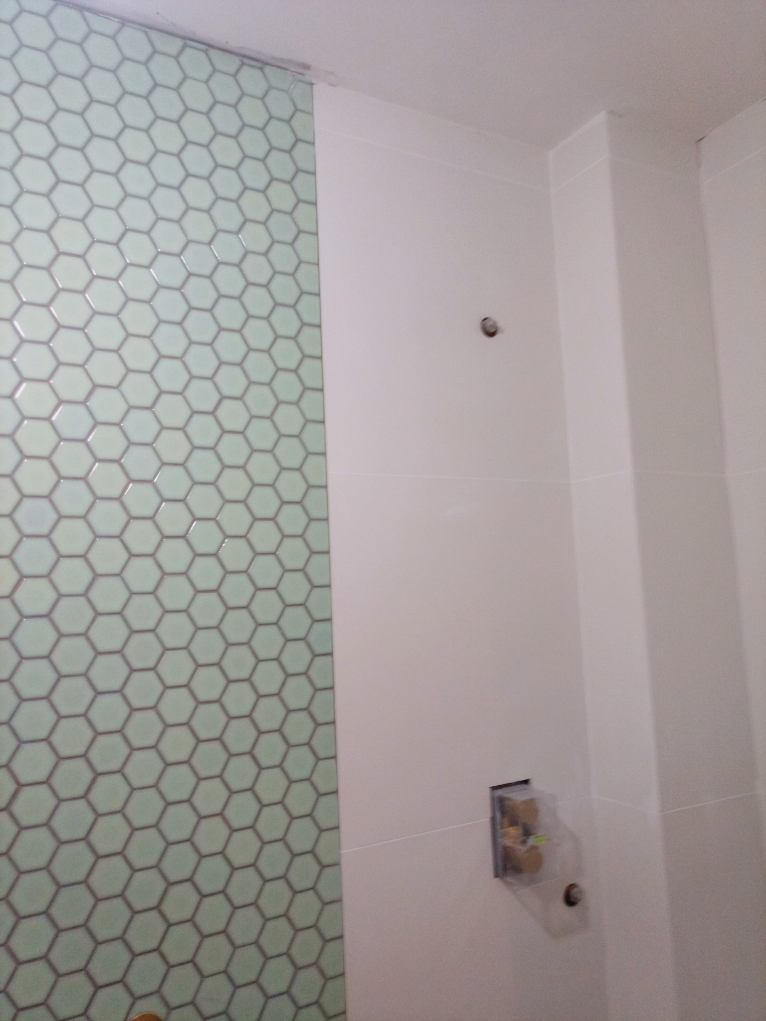 Wykończona ściana łazienki z heksagonalnymi, jasnymi płytkami. Widoczny fragment białej ściany i przygotowane miejsce na armaturę.