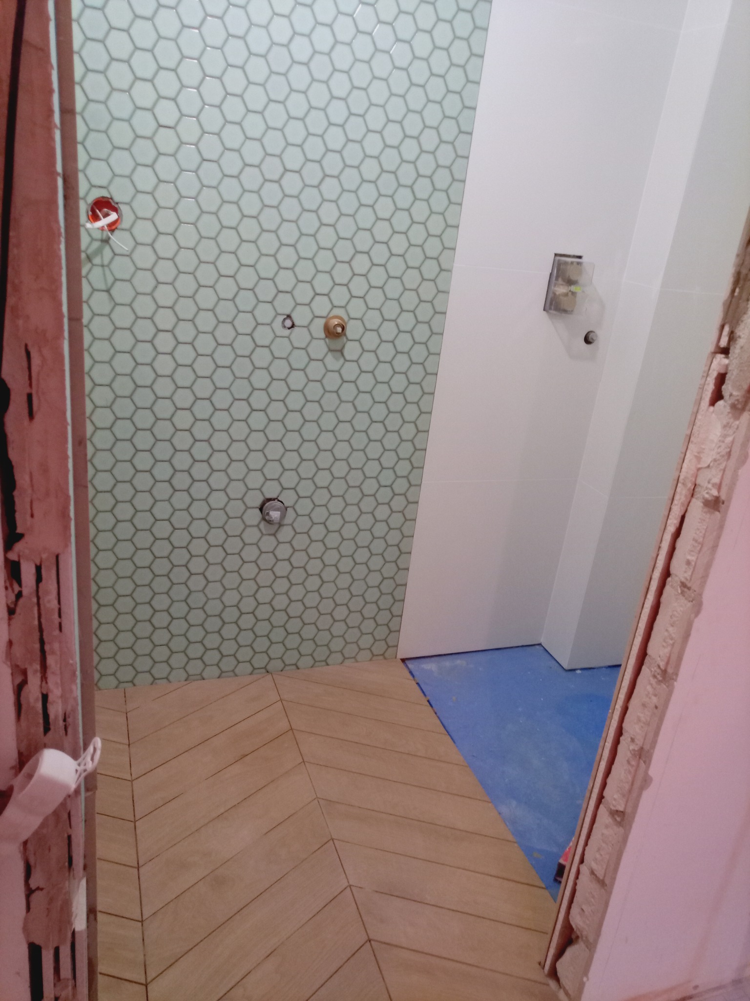 Remont łazienki: ściana z heksagonalnych płytek w odcieniach zieleni, drewnopodobna podłoga ułożona w jodełkę, widoczne instalacje i fragment surowego otworu drzwiowego.