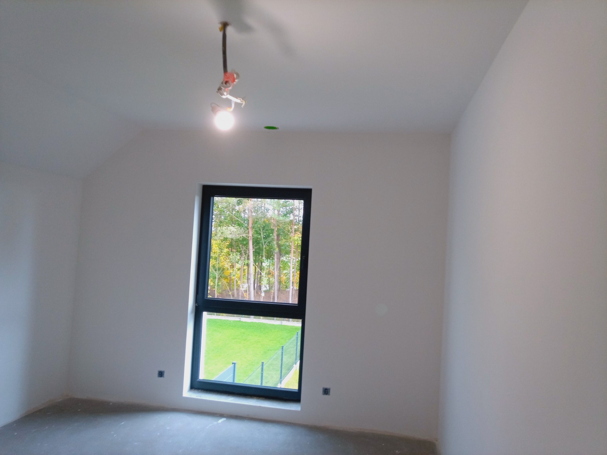 Wnętrze pokoju po malowaniu ścian na biało, widoczne czarne okno z widokiem na zieleń, surowa podłoga i wisząca lampa bez klosza.