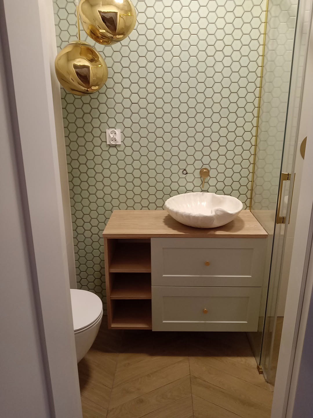 Wykończona łazienka z umywalką w kształcie muszli, szafką z szufladami i otwartymi półkami. Na ścianie heksagonalne płytki i złote lampy. Parkiet ułożony w jodełkę.