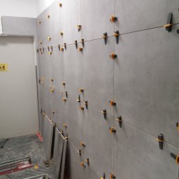 MS Construction - Układanie szarych płytek na ścianie z użyciem klipsów poziomujących i sznurka. Na podłodze widoczne narzędzia i płyty. Prace w toku, surowe wykończenie.