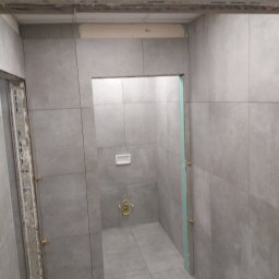 MS Construction - Szara, wykafelkowana łazienka w trakcie remontu. Widoczne płytki, elementy instalacji, listwy montażowe i poziomica. W tle toaleta podtynkowa z żółtymi elementami.