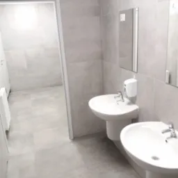 Jasna łazienka z dwoma umywalkami na postumentach, duże szare płytki na ścianach i podłodze, białe drzwi w tle. Minimalistyczny design.