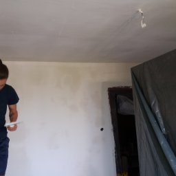 MS CONSTRUCTIONS MICHAŁ STUDZIŃSKI - Mężczyzna szpachluje ścianę w pomieszczeniu, widoczne narzędzia i fragment zakrytego folią pomieszczenia. Ściana częściowo wyszpachlowana.