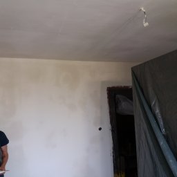 MS CONSTRUCTIONS MICHAŁ STUDZIŃSKI - Ściana po szpachlowaniu, widoczne niedoskonałości przed malowaniem. Zabezpieczone drzwi folią malarską. Fragment osoby z notatnikiem w lewym dolnym rogu kadru.