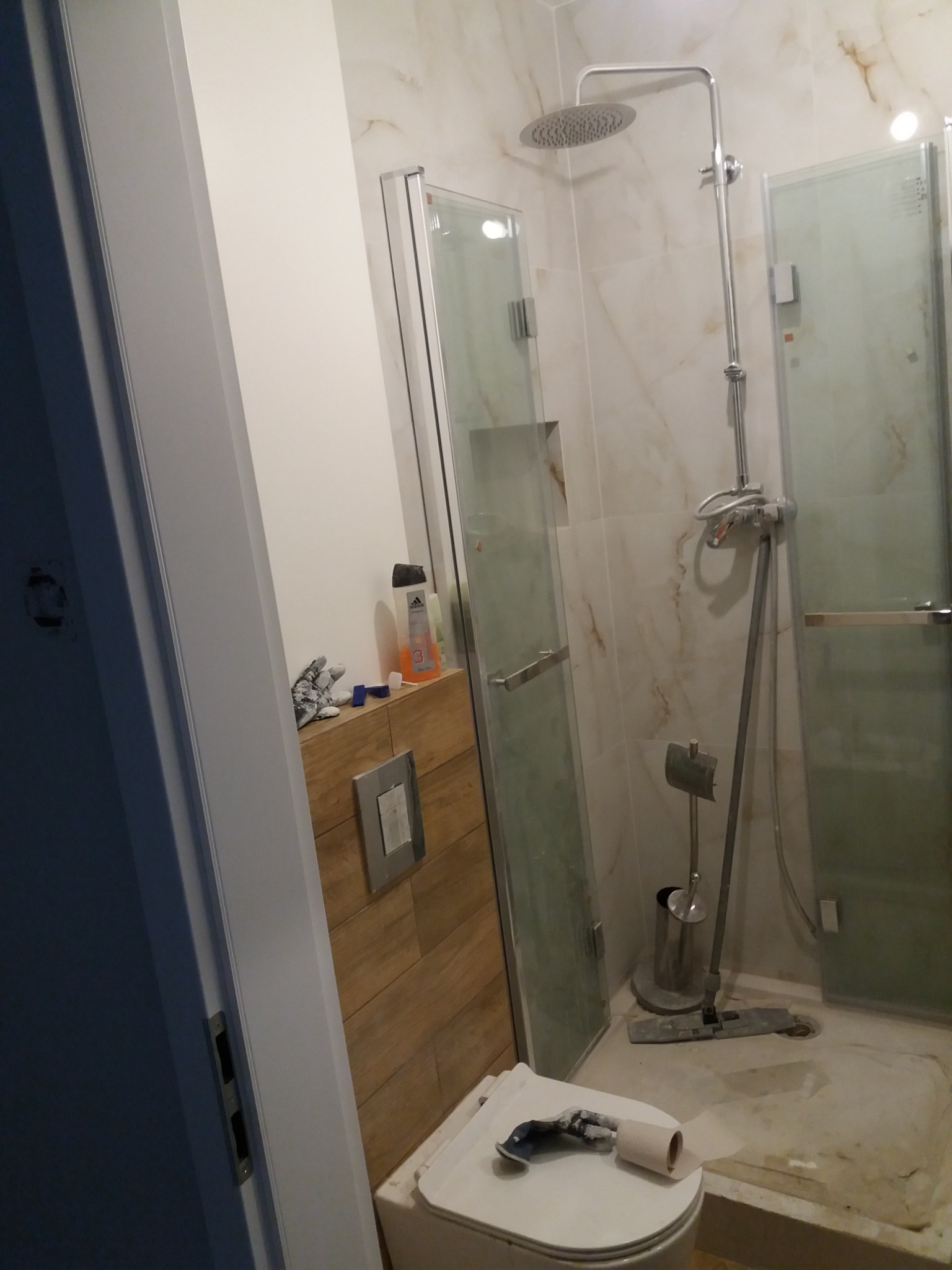 Wykończona łazienka z kabiną prysznicową, marmurowymi płytkami i drewnianą zabudową umywalki. Na sedesie leżą narzędzia i papier toaletowy.