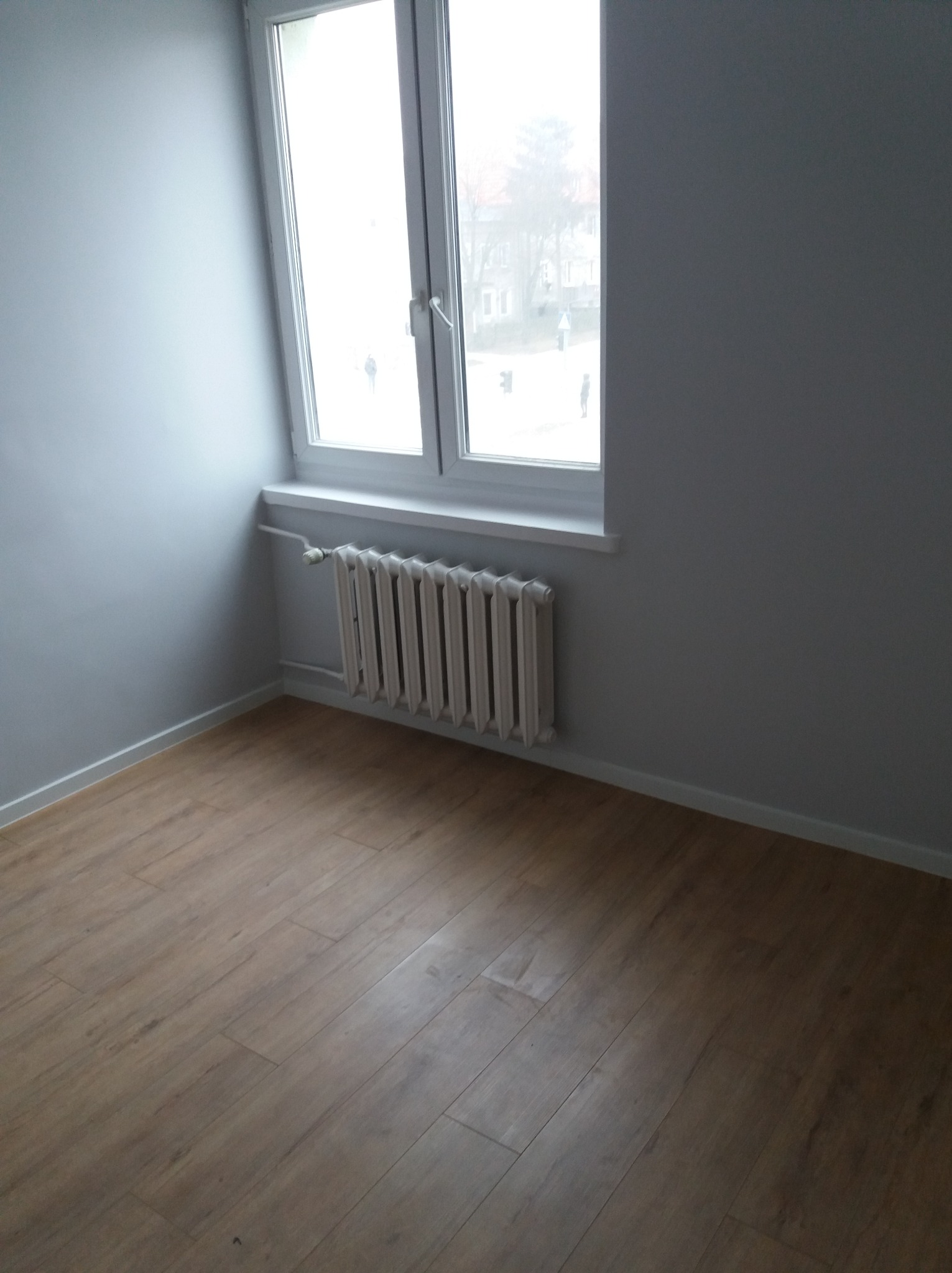 Jasny pokój po remoncie: panele podłogowe, szare ściany, białe okno z widokiem na zewnątrz i grzejnik. Minimalistyczne wykończenie wnętrza.
