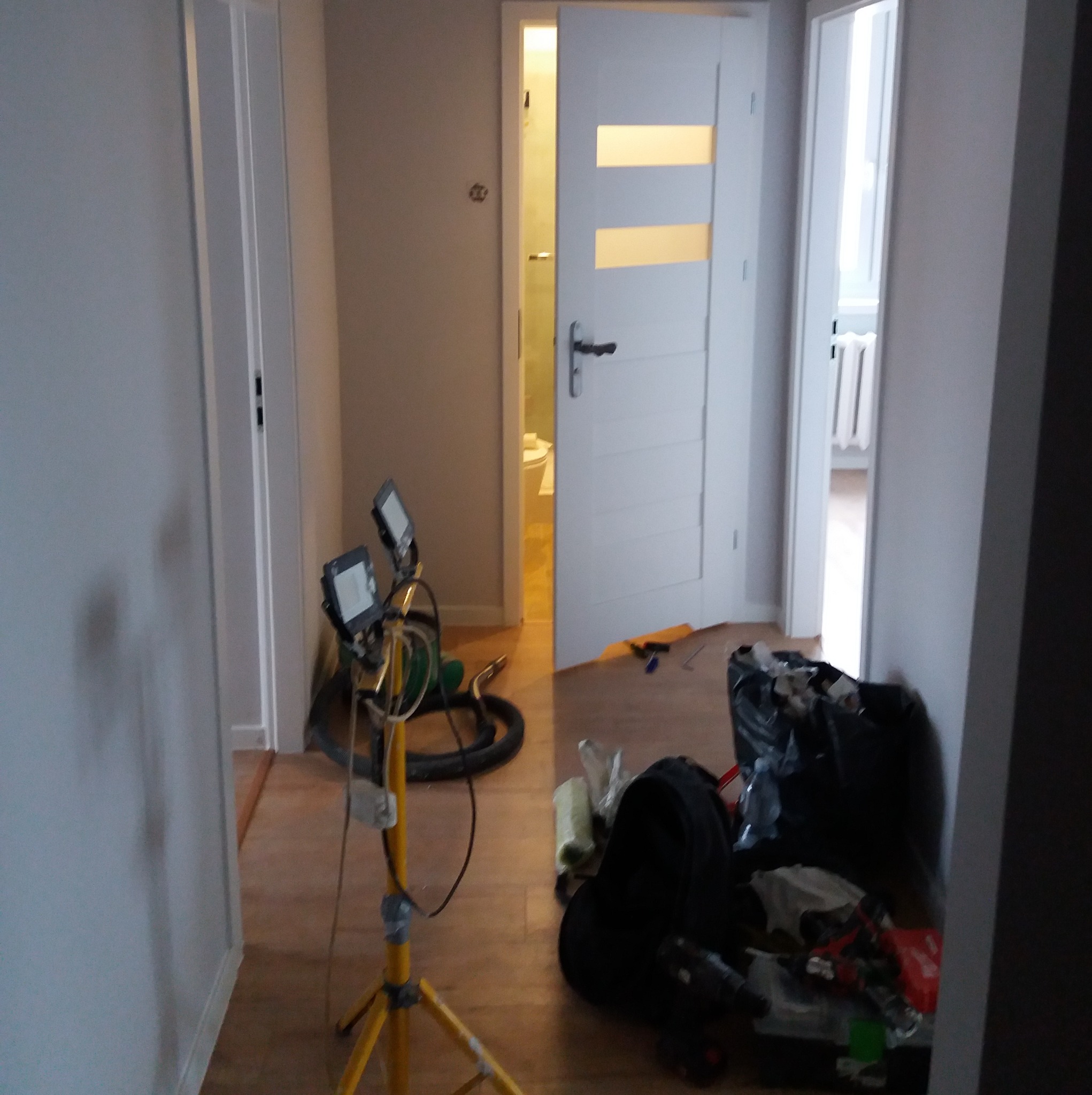 Wnętrze mieszkania w trakcie prac wykończeniowych: widoczne otwarte drzwi z przeszkleniami, lampa budowlana, podłoga i ściany po malowaniu. W tle widoczna łazienka.