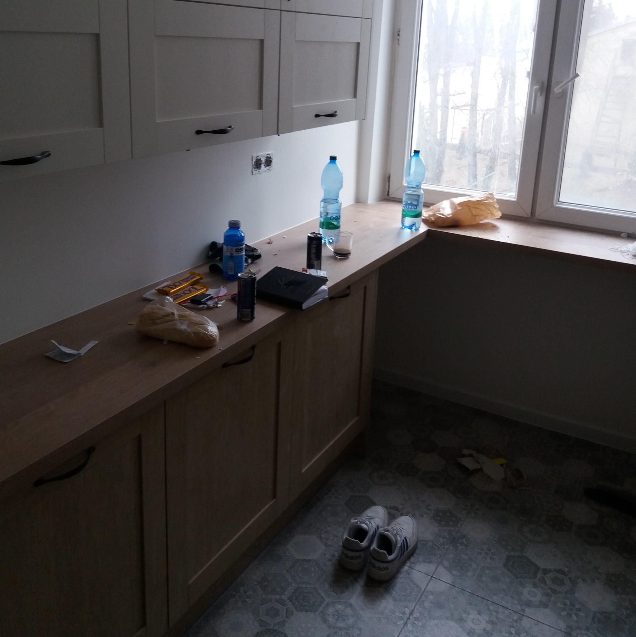 Kuchnia w trakcie remontu: widoczne białe szafki, blat z drobnymi przedmiotami, okno i podłoga z szarymi płytkami. Rozpoczęty etap prac, bałagan.