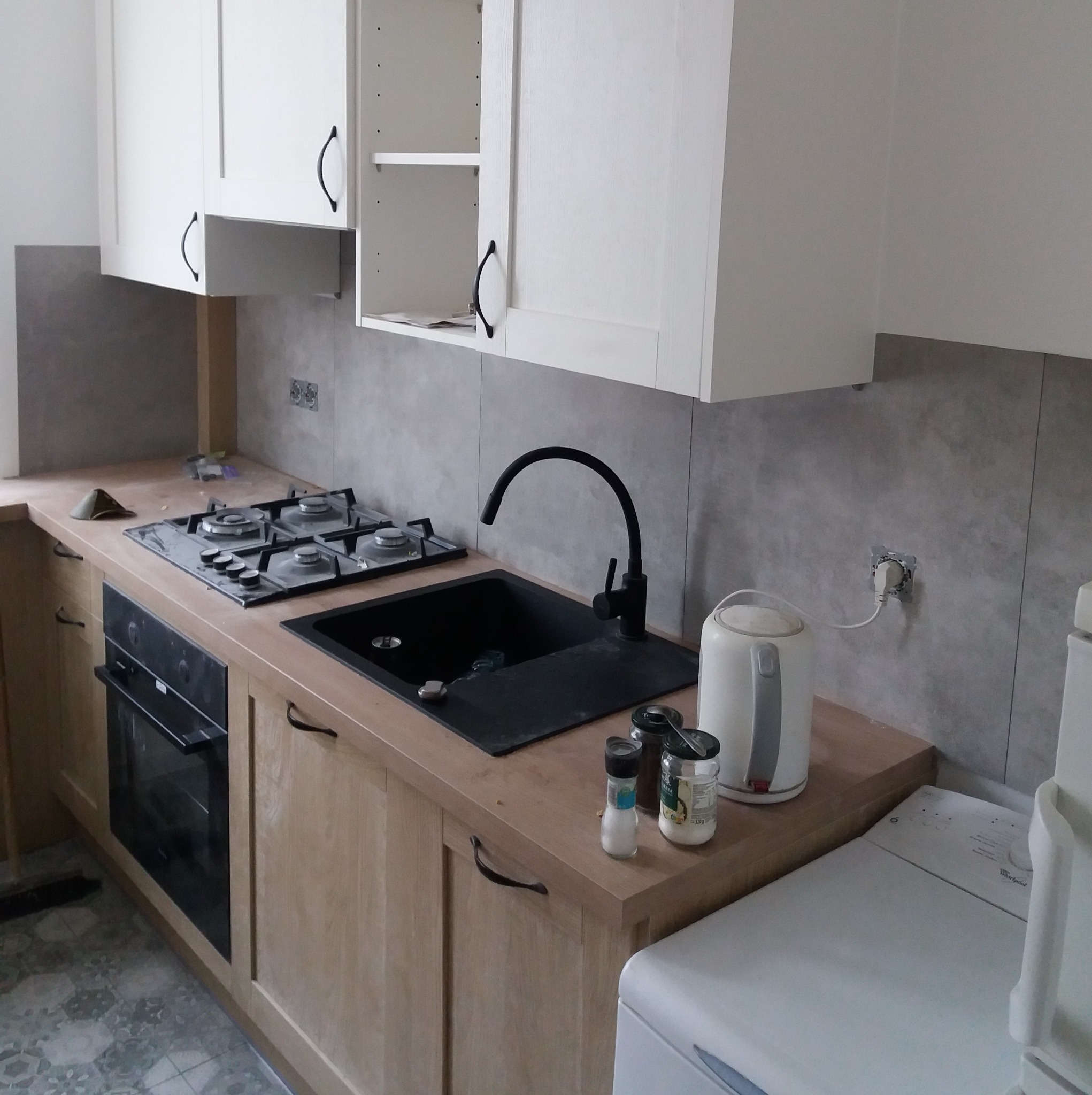 Kuchnia w trakcie montażu: jasne drewniane szafki dolne z czarnymi uchwytami, białe szafki górne, czarny zlew i bateria, płyta gazowa. Widoczny fragment pralki.