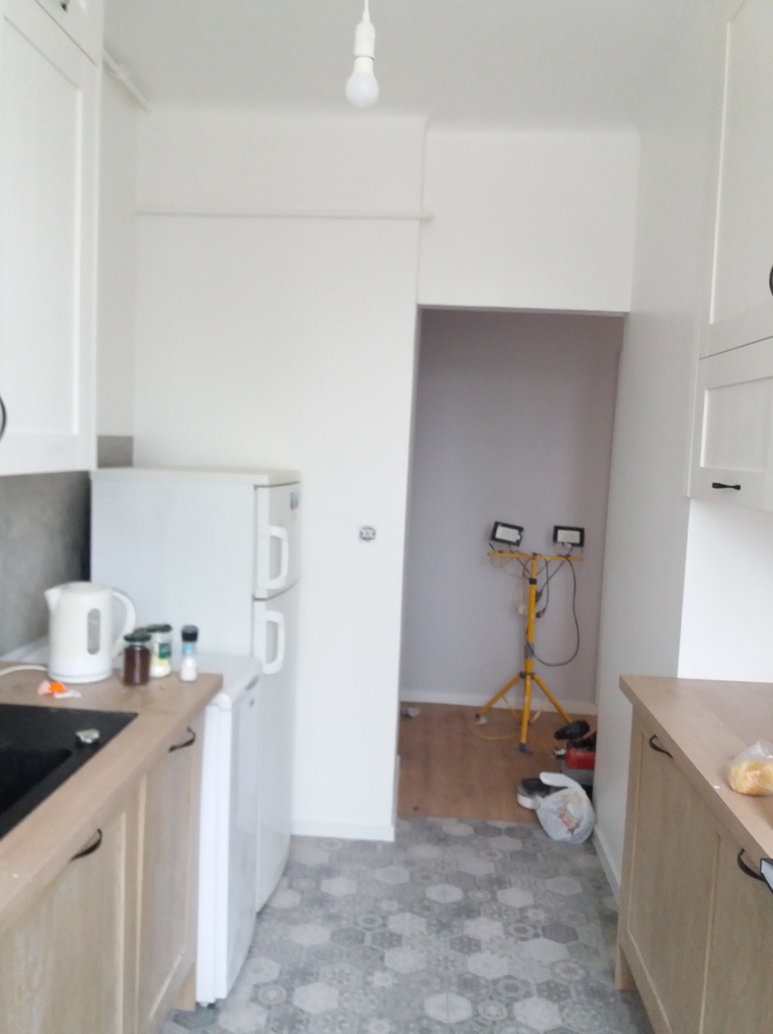 Wnętrze kuchni w trakcie remontu. Widoczne nowe meble, lodówka, podłoga w heksagonalne wzory i lampy robocze. Ściany przygotowane do malowania.