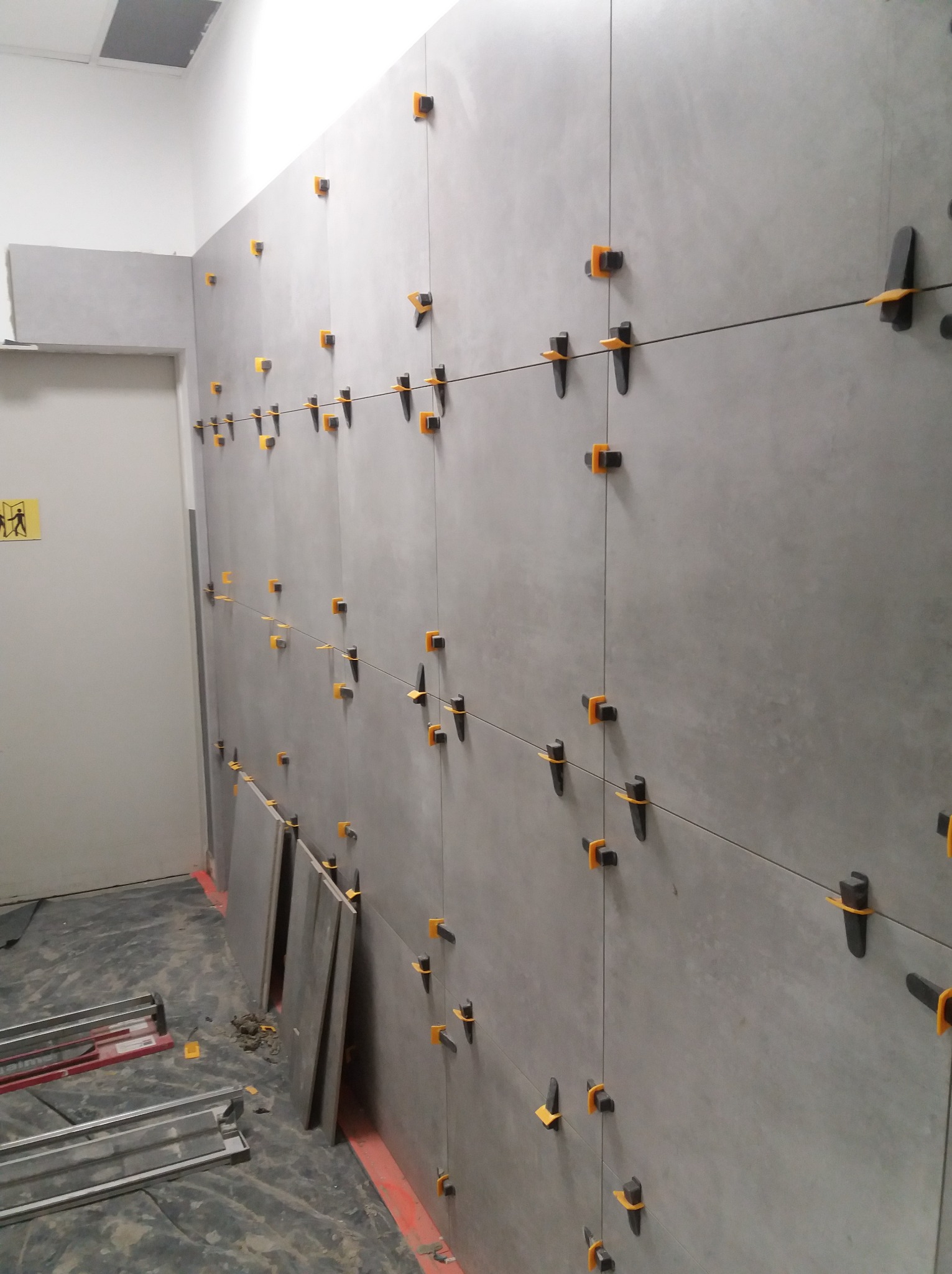 Układanie szarych płytek na ścianie z użyciem klipsów poziomujących i sznurka. Na podłodze widoczne narzędzia i płyty. Prace w toku, surowe wykończenie.