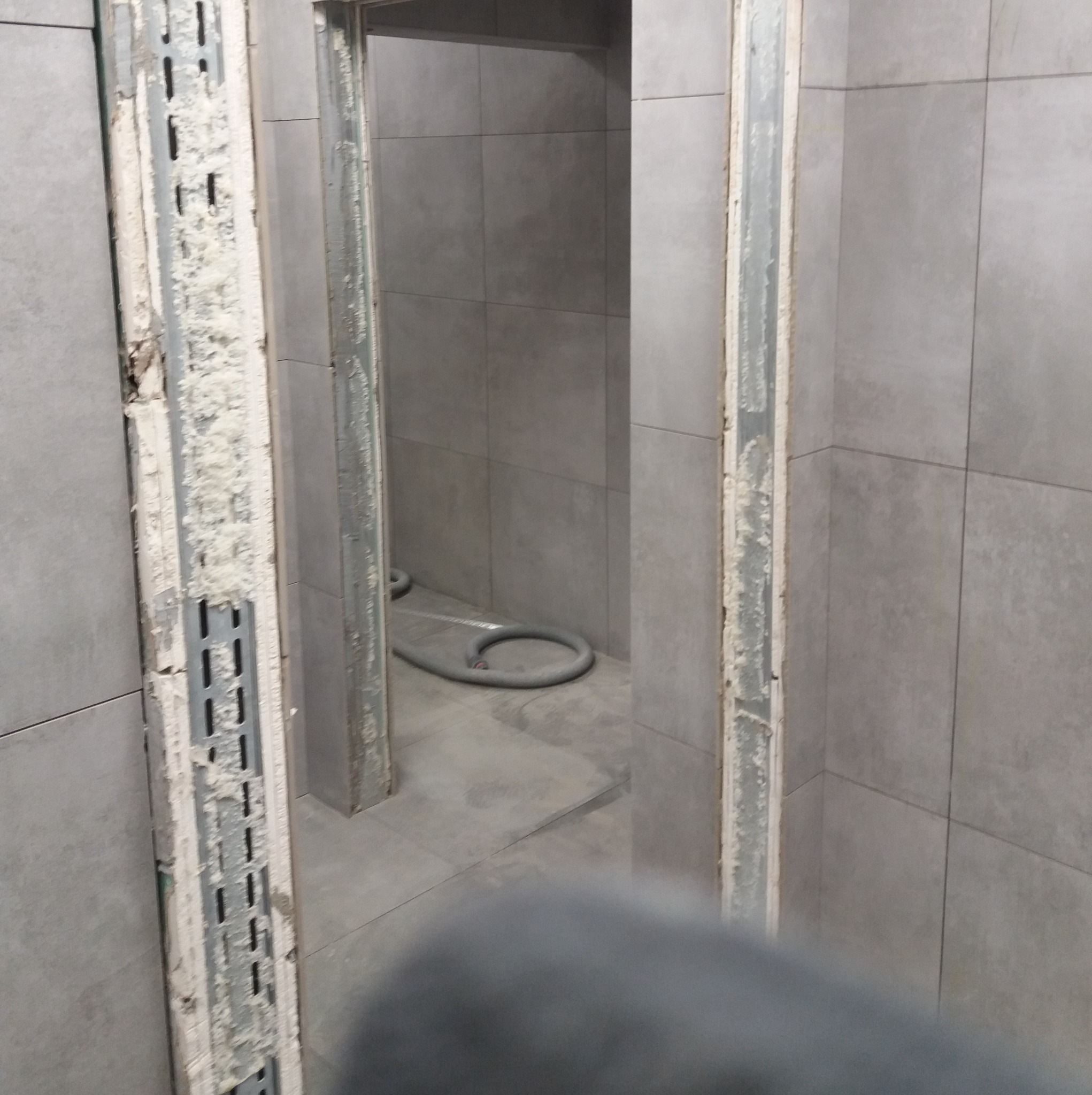 Wnętrze łazienki w trakcie remontu. Widoczne szare płytki, metalowe profile montażowe przy ościeżnicy i fragment węża kanalizacyjnego. Prace wykończeniowe w toku.