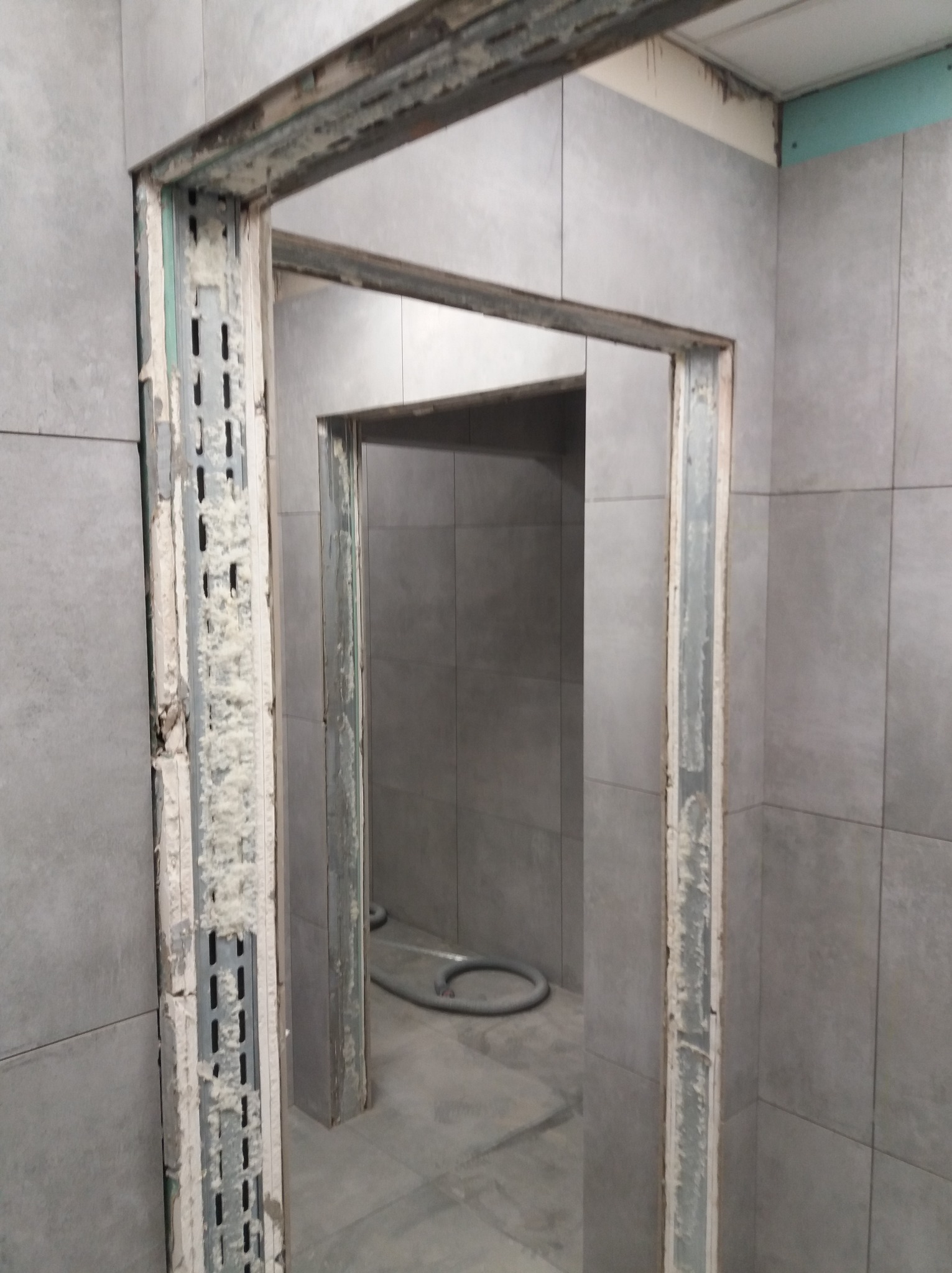 Łazienka w trakcie remontu: widoczne metalowe profile ościeżnicy z pianką montażową, szare płytki na ścianach i podłodze. W tle widoczny fragment węża.