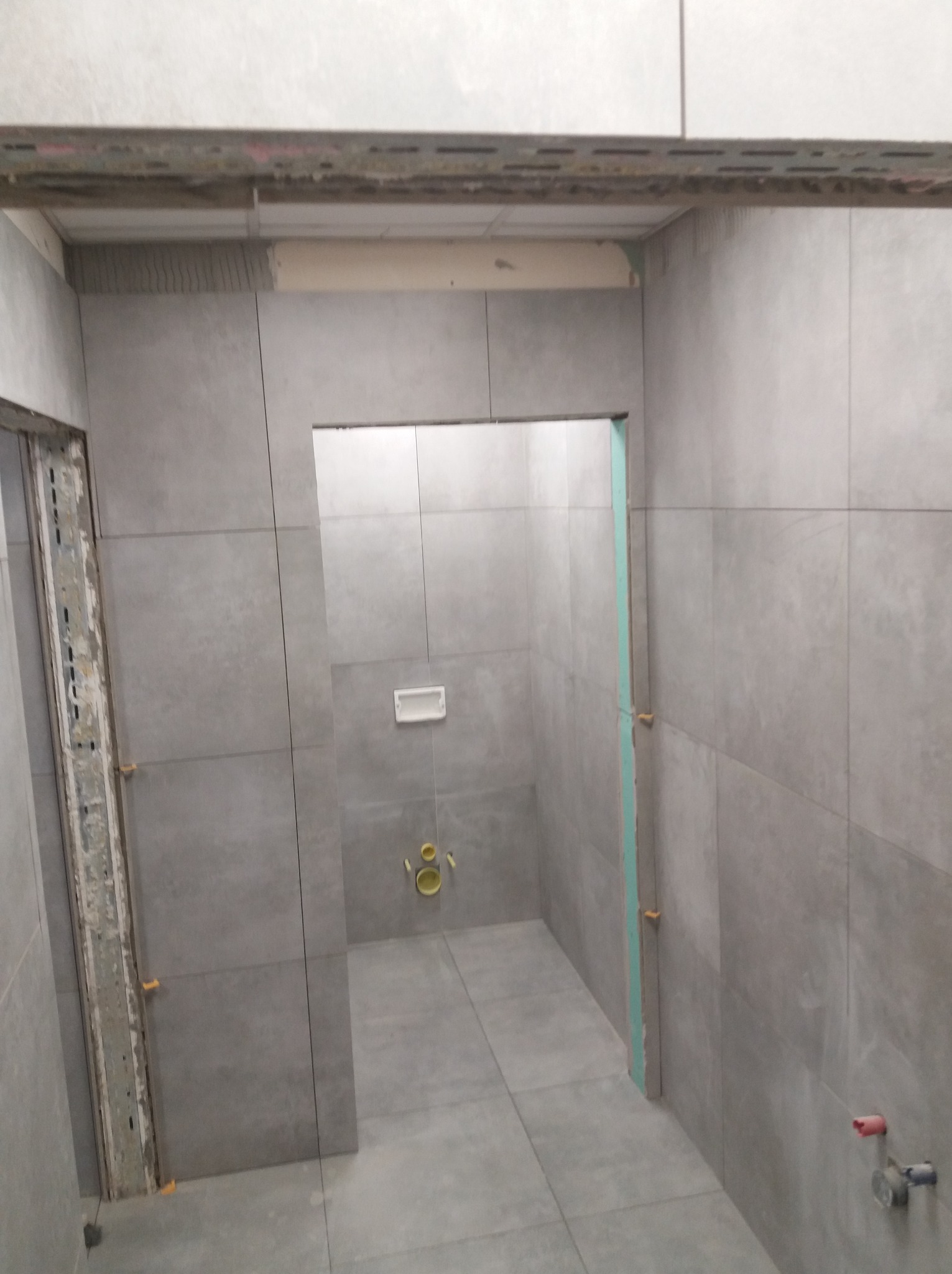 Szara, wykafelkowana łazienka w trakcie remontu. Widoczne płytki, elementy instalacji, listwy montażowe i poziomica. W tle toaleta podtynkowa z żółtymi elementami.