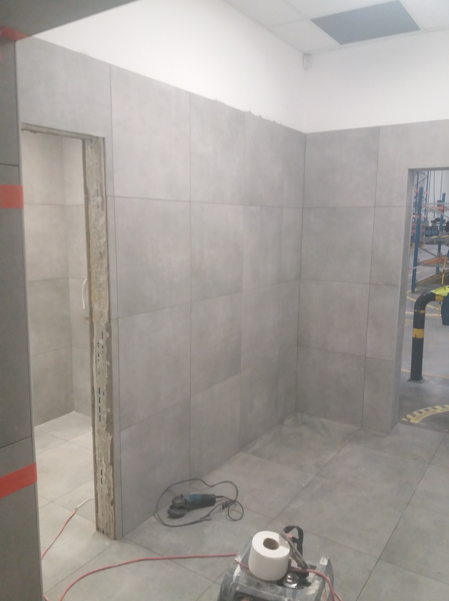 Wnętrze w trakcie remontu: szare płytki na ścianach i podłodze, widoczne narzędzia - szlifierka i maszyna do cięcia płytek, otwór drzwiowy bez ościeżnicy.