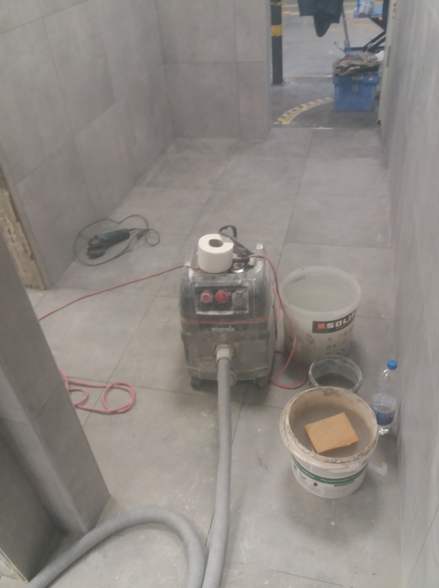 Wnętrze łazienki w trakcie prac. Widoczny odkurzacz piorący, wiadra z wodą i gąbką, szlifierka kątowa i rolka papieru. Szare płytki na ścianach i podłodze.