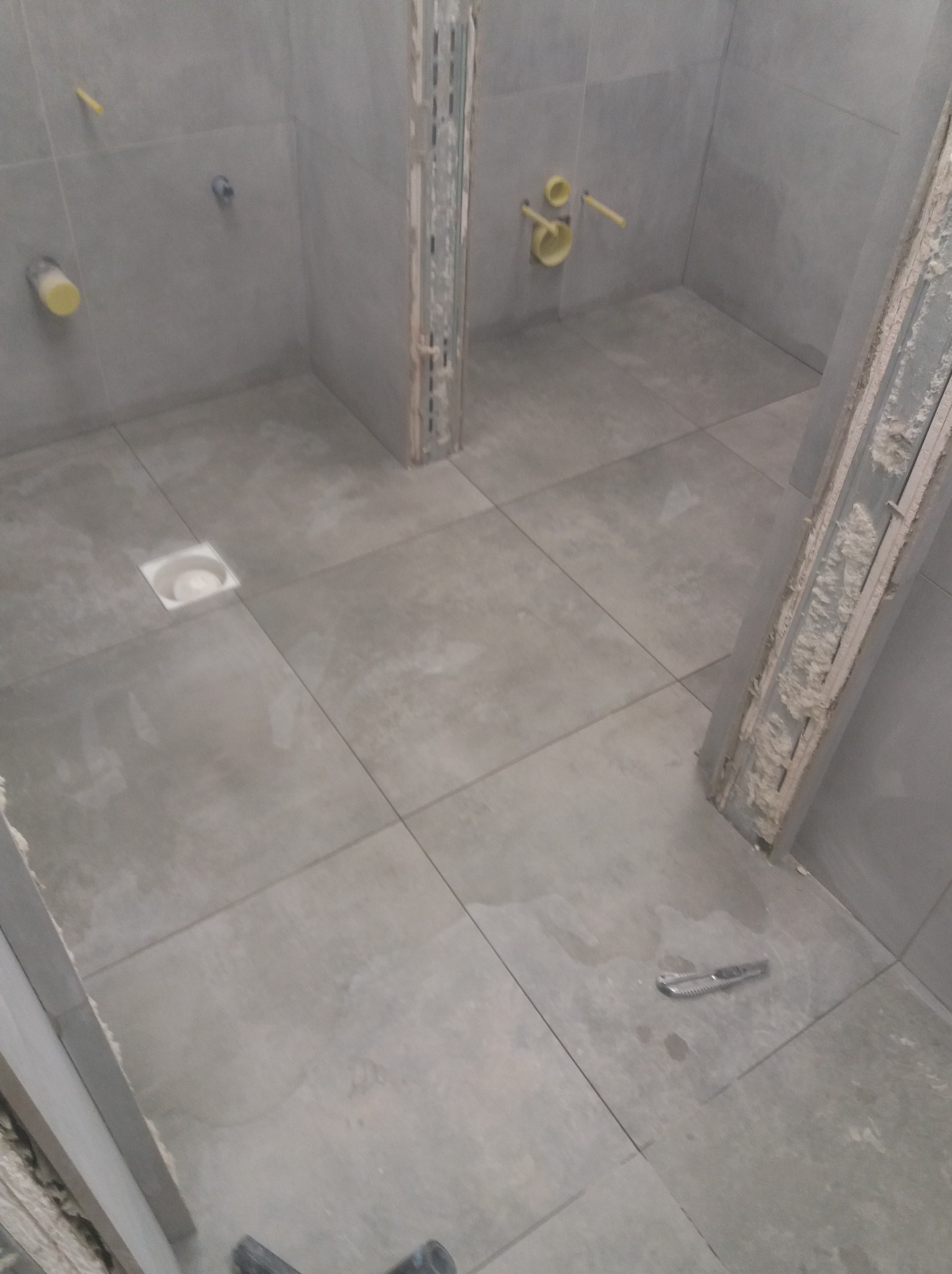 Szare płytki w łazience w trakcie remontu, widoczne instalacje i narzędzie. Ujęcie z góry, ukazujące postęp prac.