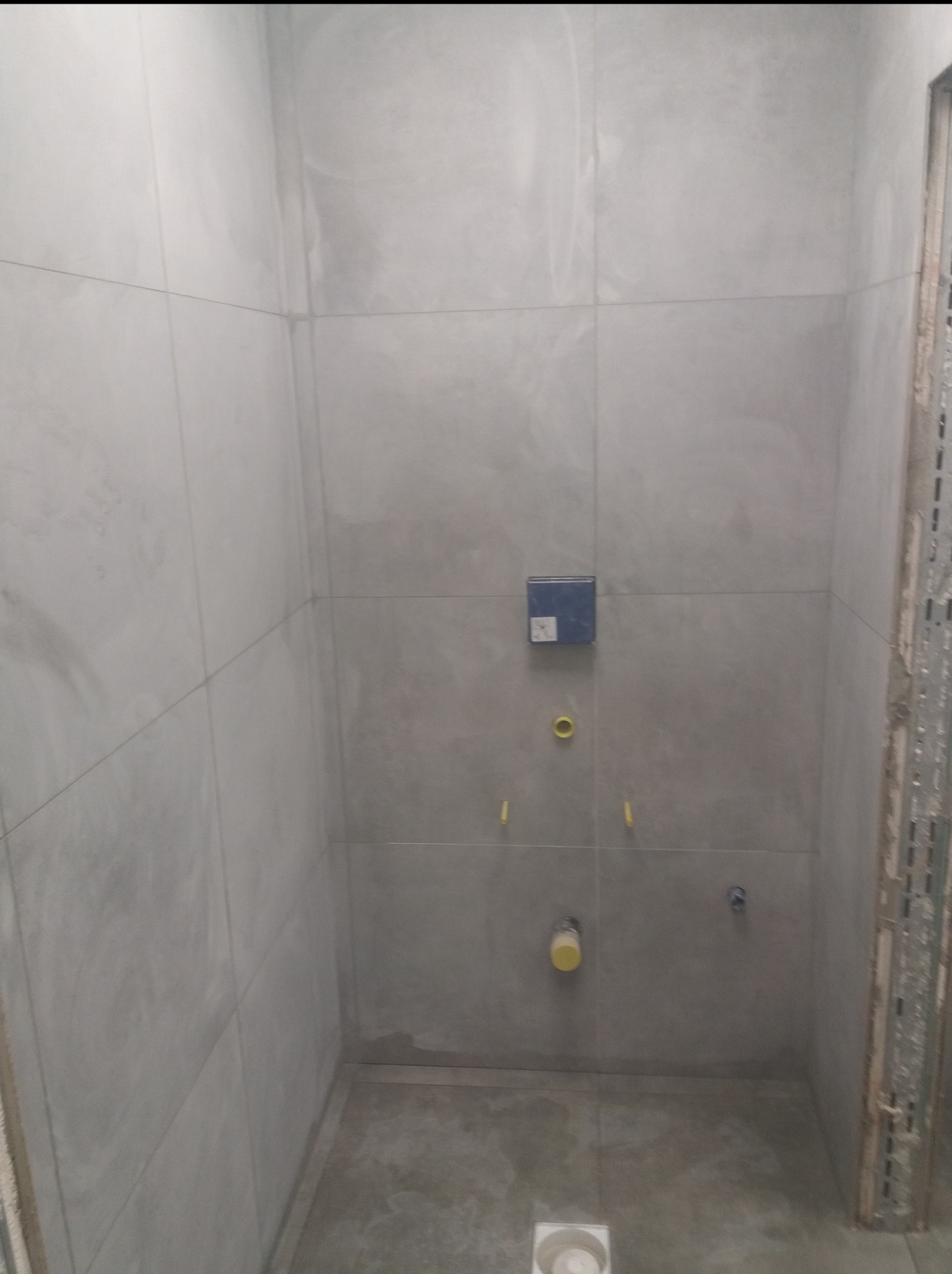 Ściana wyłożona szarymi płytkami w łazience w trakcie remontu. Widoczne otwory na armaturę i stelaż podtynkowy. W tle fragment metalowej konstrukcji.