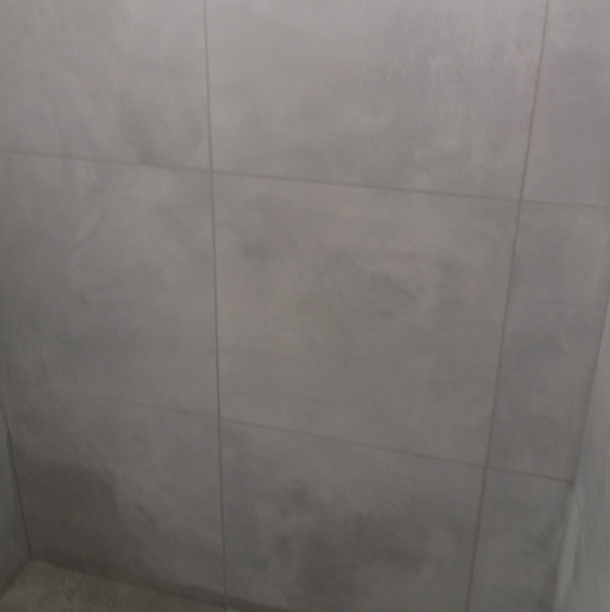 Wykończona ściana łazienki z szarymi, matowymi płytkami ceramicznymi ułożonymi w regularny wzór. Widoczna fuga w kolorze zbliżonym do płytek.