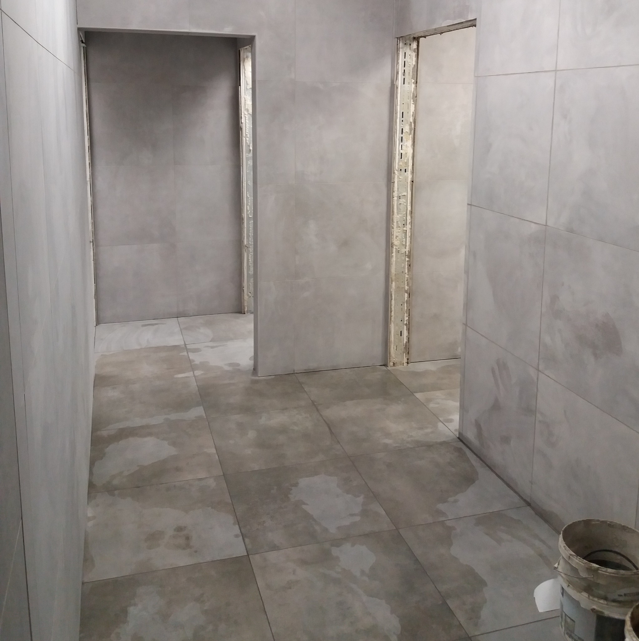 Szare płytki podłogowe i ścienne w trakcie układania, widoczne dwa otwory drzwiowe bez ościeżnic. Wiadro z zaprawą w rogu. Wnętrze w trakcie prac wykończeniowych.