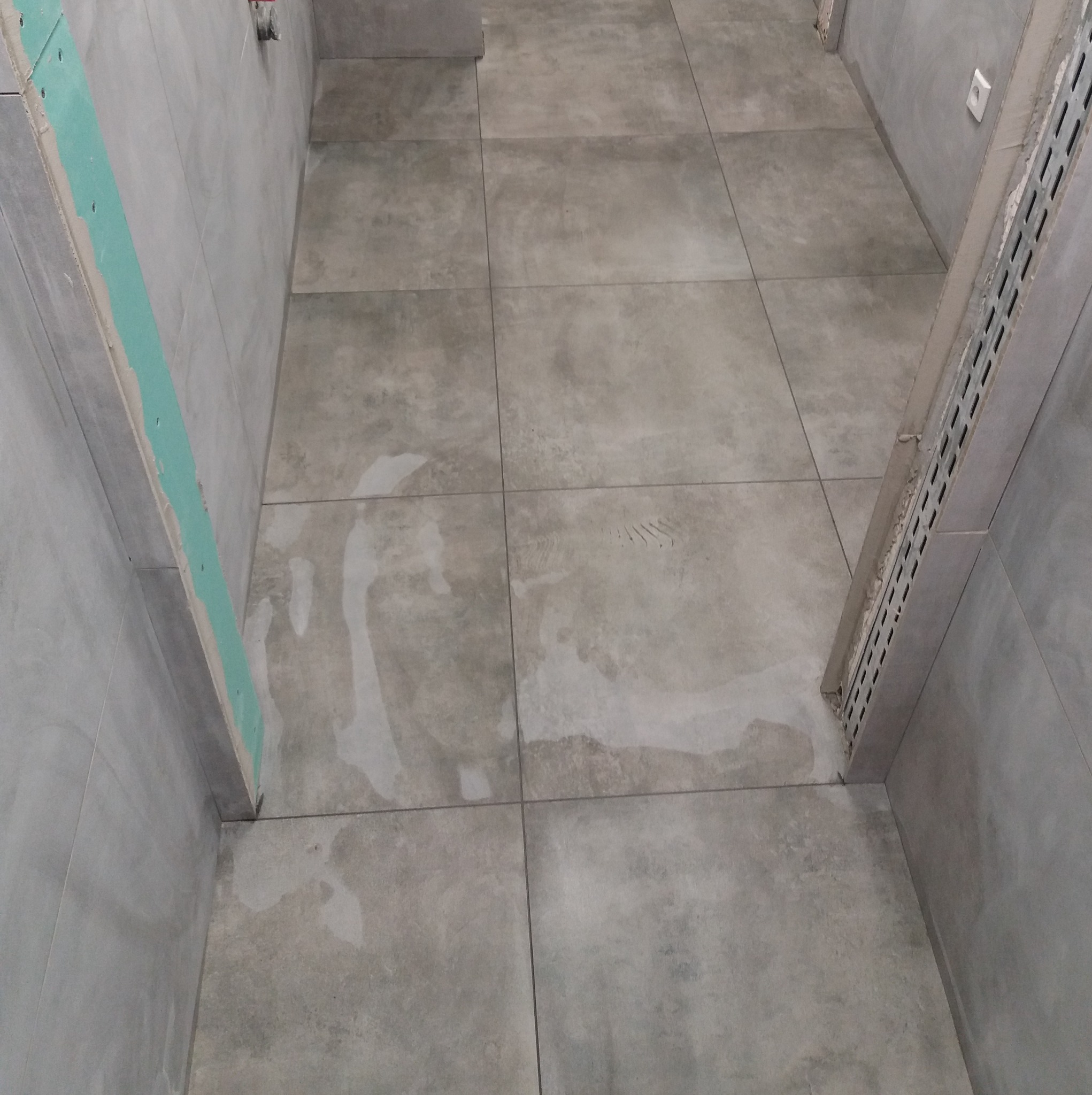 Szare płytki podłogowe w trakcie układania w łazience, widoczne ślady po kleju i fragment ściany z profilem montażowym. Surowy, minimalistyczny styl wykończenia.