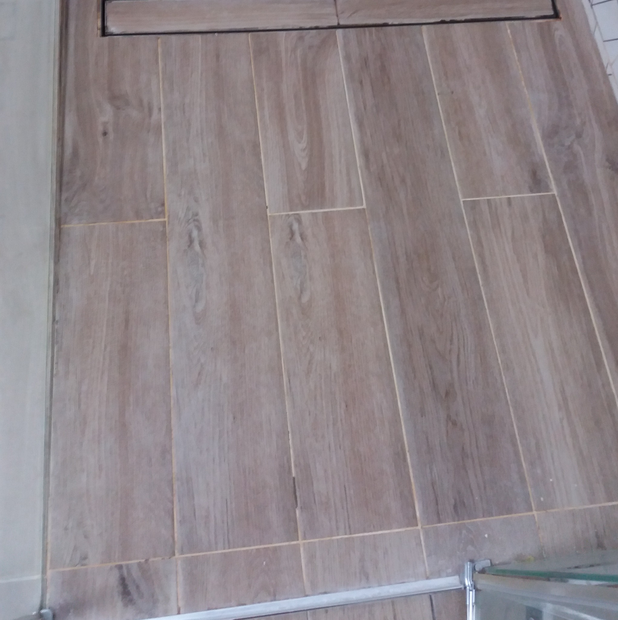 Ułożone płytki podłogowe imitujące drewno w łazience, z widocznym odpływem liniowym i fragmentem kabiny prysznicowej. Jasne fugi podkreślają wzór.