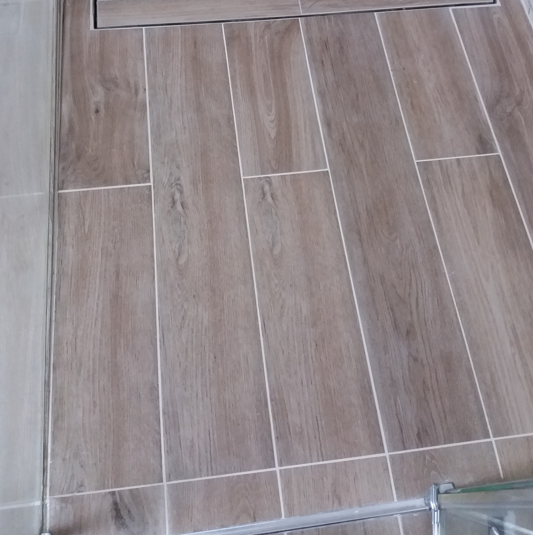 Ułożona podłoga z płytek imitujących drewno w łazience, z widocznym odpływem liniowym i fragmentem szklanej kabiny prysznicowej. Jasne fugi podkreślają układ.