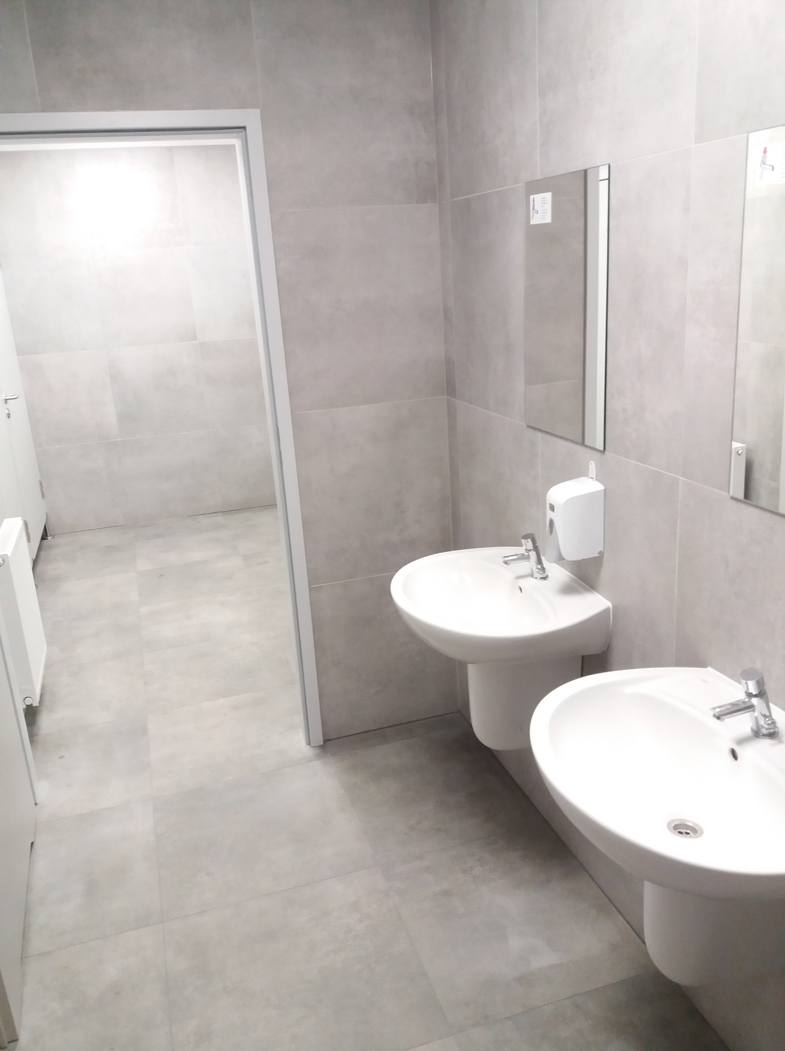 Jasna łazienka z dwoma umywalkami na postumentach, duże szare płytki na ścianach i podłodze, białe drzwi w tle. Minimalistyczny design.