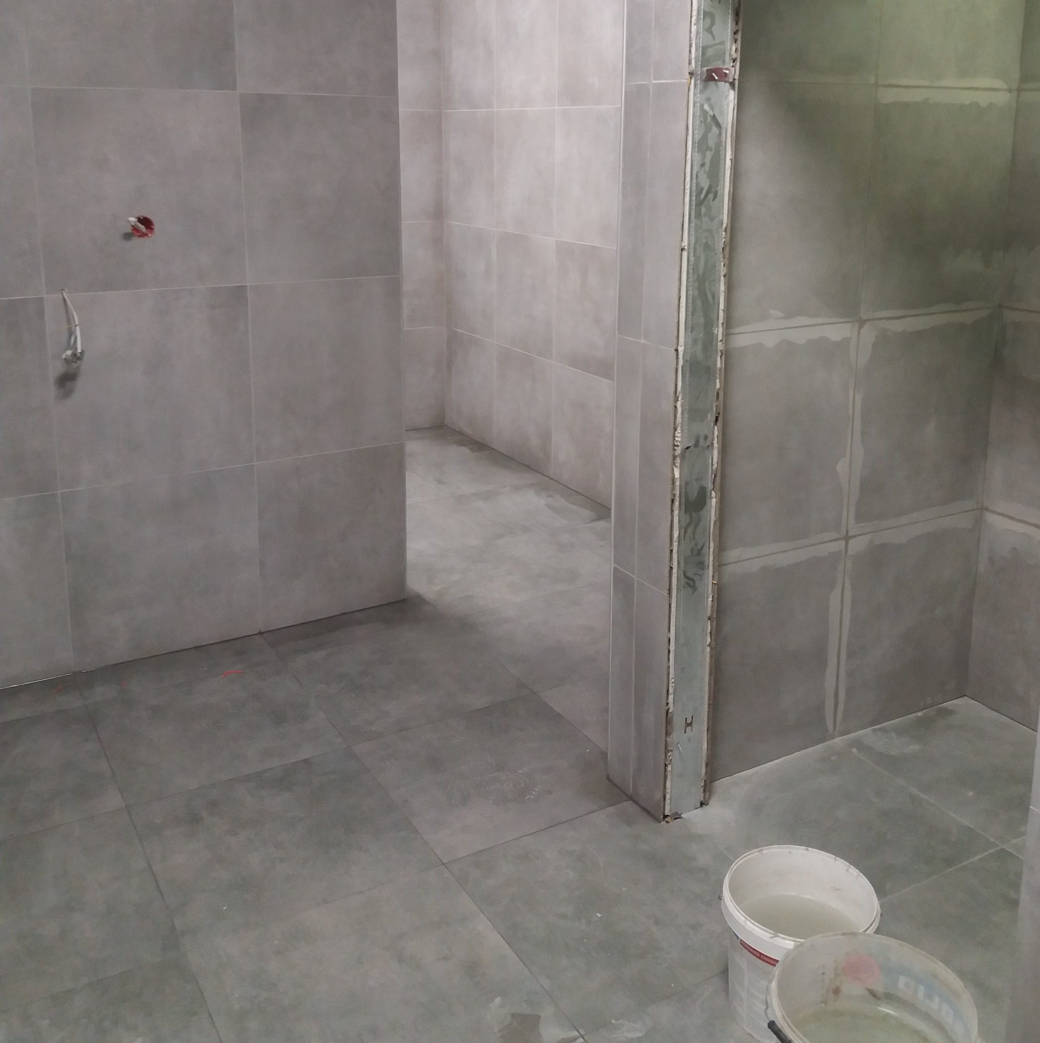 Szare płytki w trakcie montażu w łazience. Widoczne przygotowane ściany, podłoga i fragment futryny. Dwa wiadra z wodą w rogu pomieszczenia.