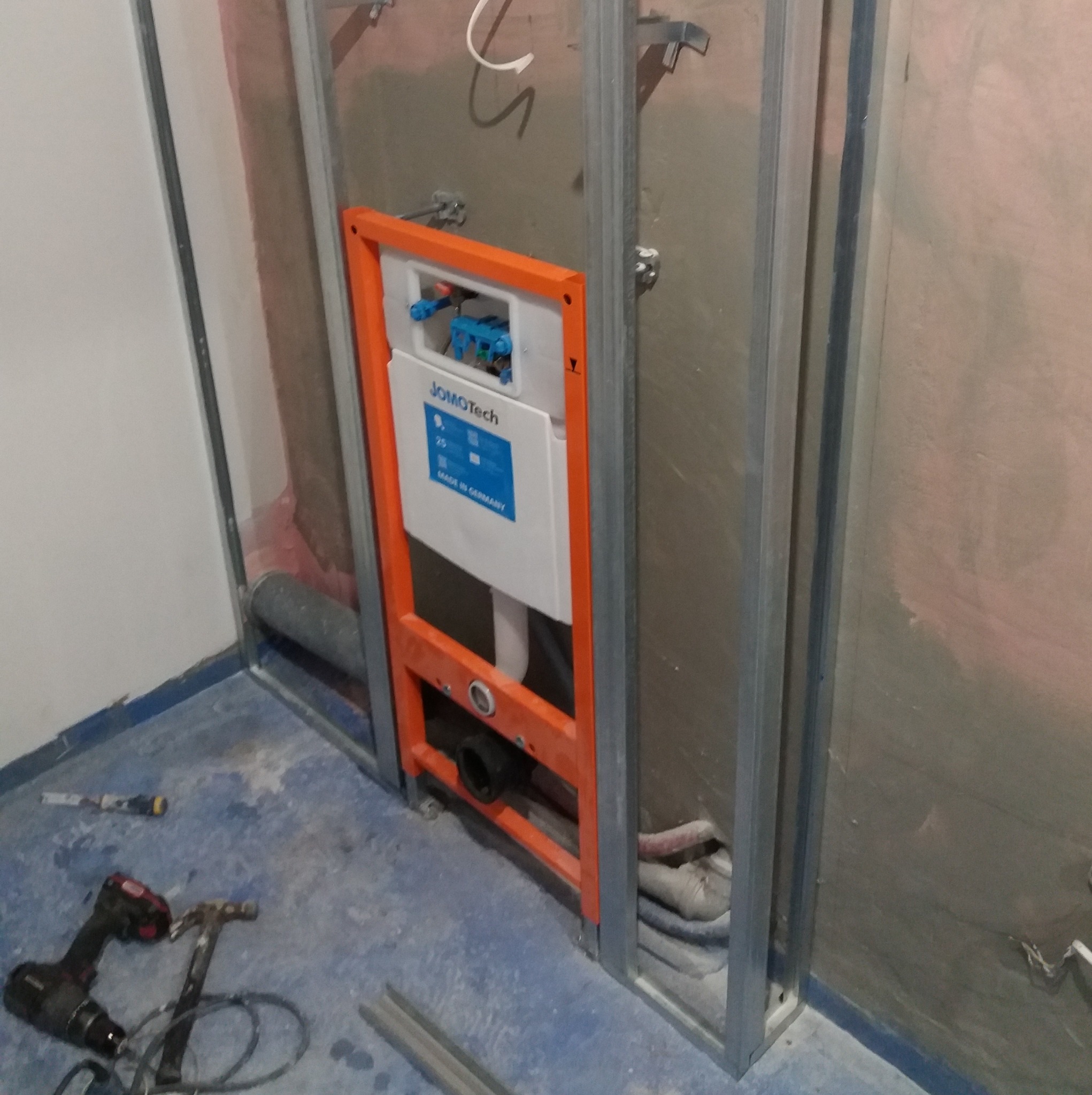 Stelaż podtynkowy Jomotech z pomarańczową ramą, w trakcie montażu w metalowej konstrukcji szkieletowej, widoczne narzędzia na niebieskiej posadzce.