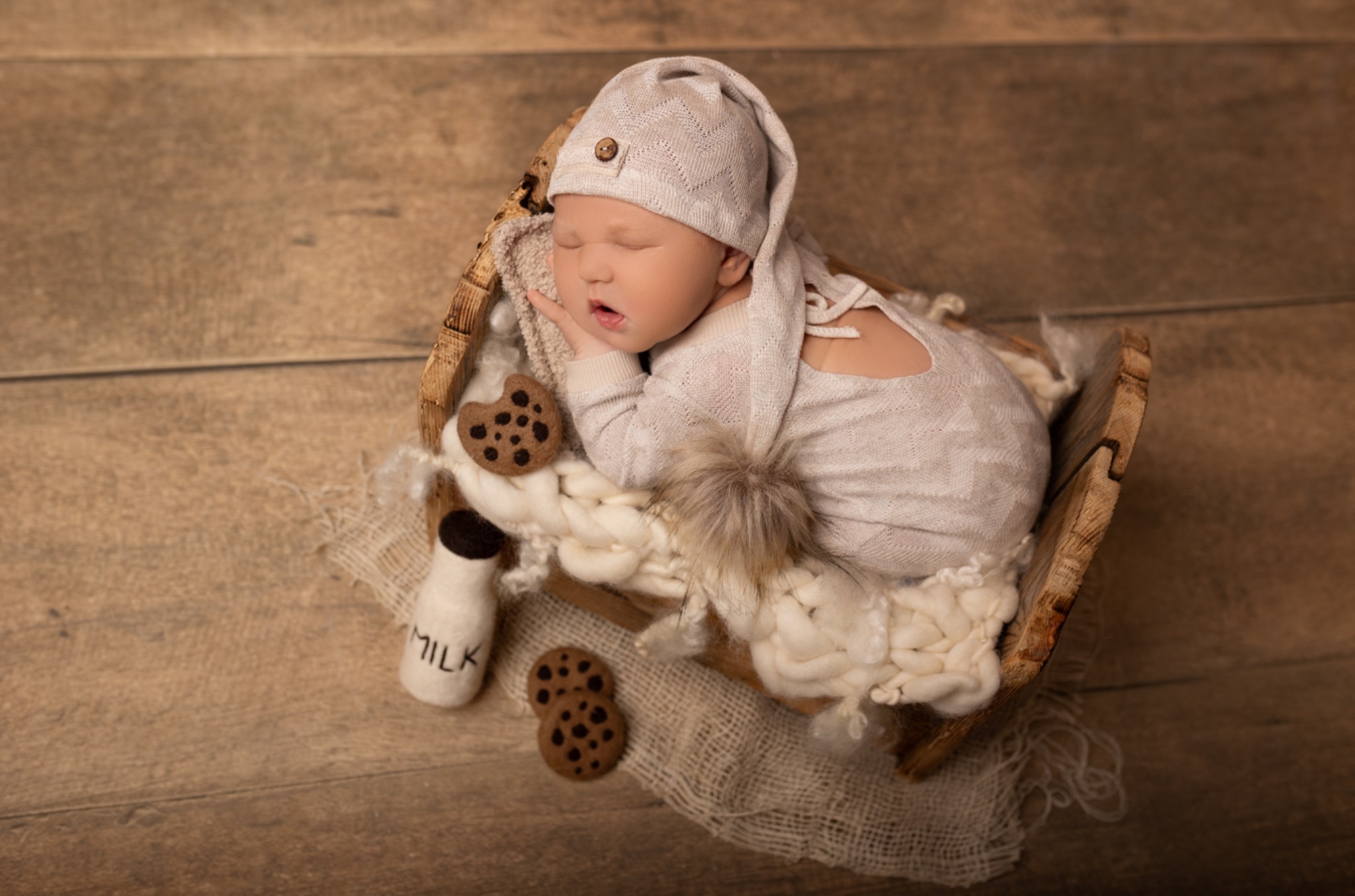 Śpiący noworodek w beżowym stroju i czapce, ułożony w drewnianym koszyku z wełnianym kocykiem, obok filcowe ciasteczka i butelka mleka na drewnianym tle.