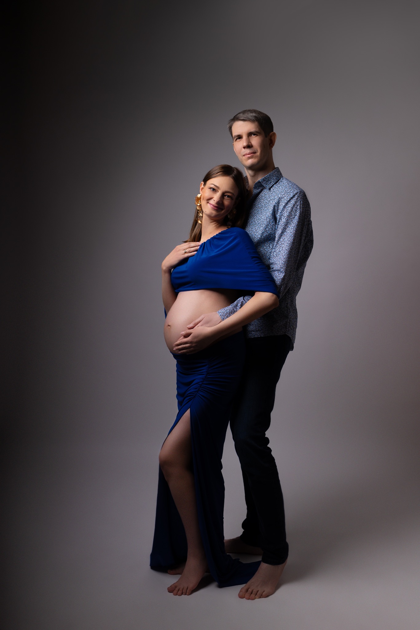 Sesja ciążowa w studio: przyszła mama w długiej, kobaltowej sukni z rozcięciem, partner obejmuje ją czule, oboje uśmiechnięci, stonowane tło.