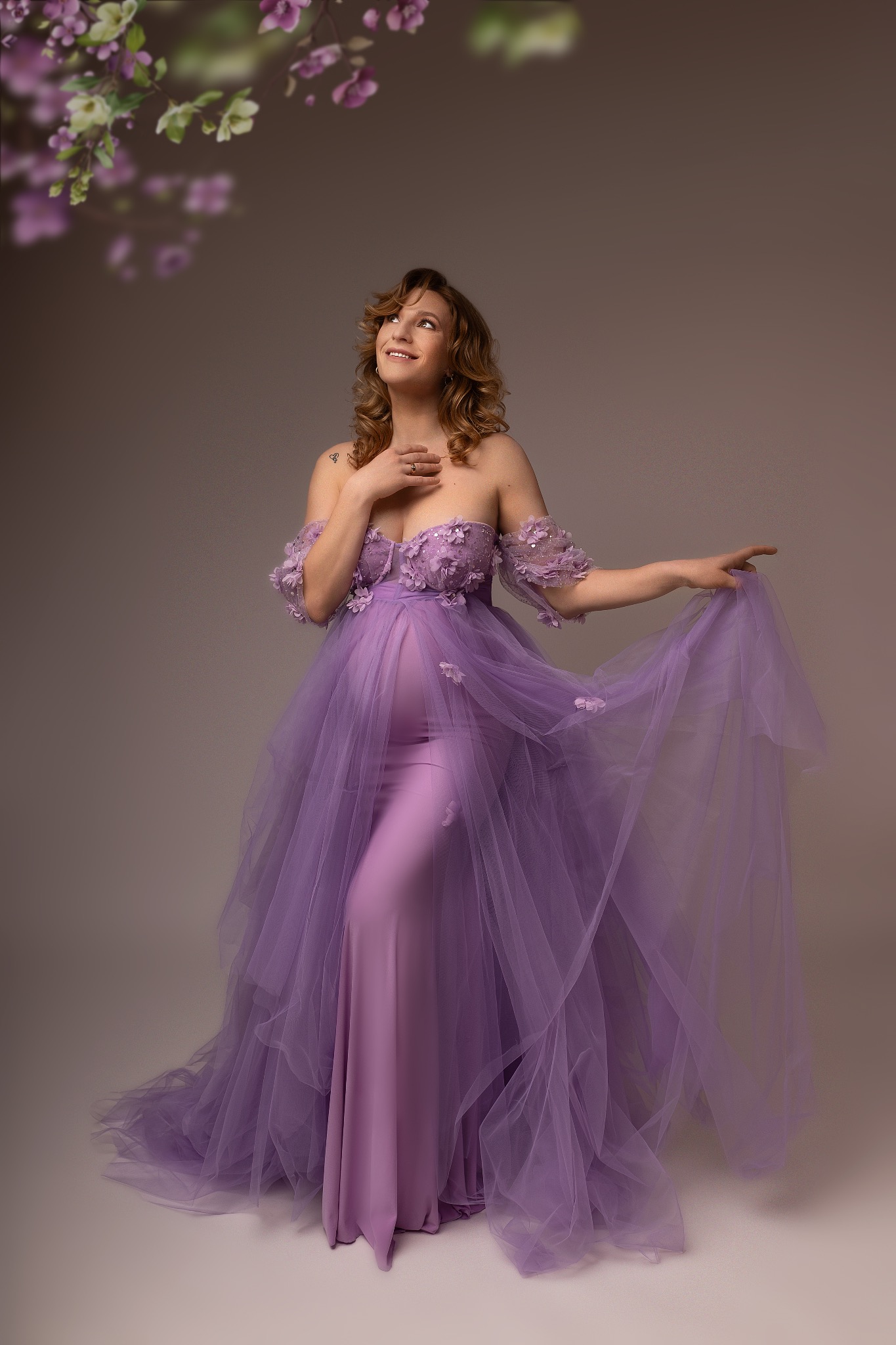 Kobieta w ciąży w fioletowej sukni z kwiatami, patrzy w górę na kwiaty. Romantyczna sesja w studiu z delikatnym oświetleniem i subtelnym tłem.