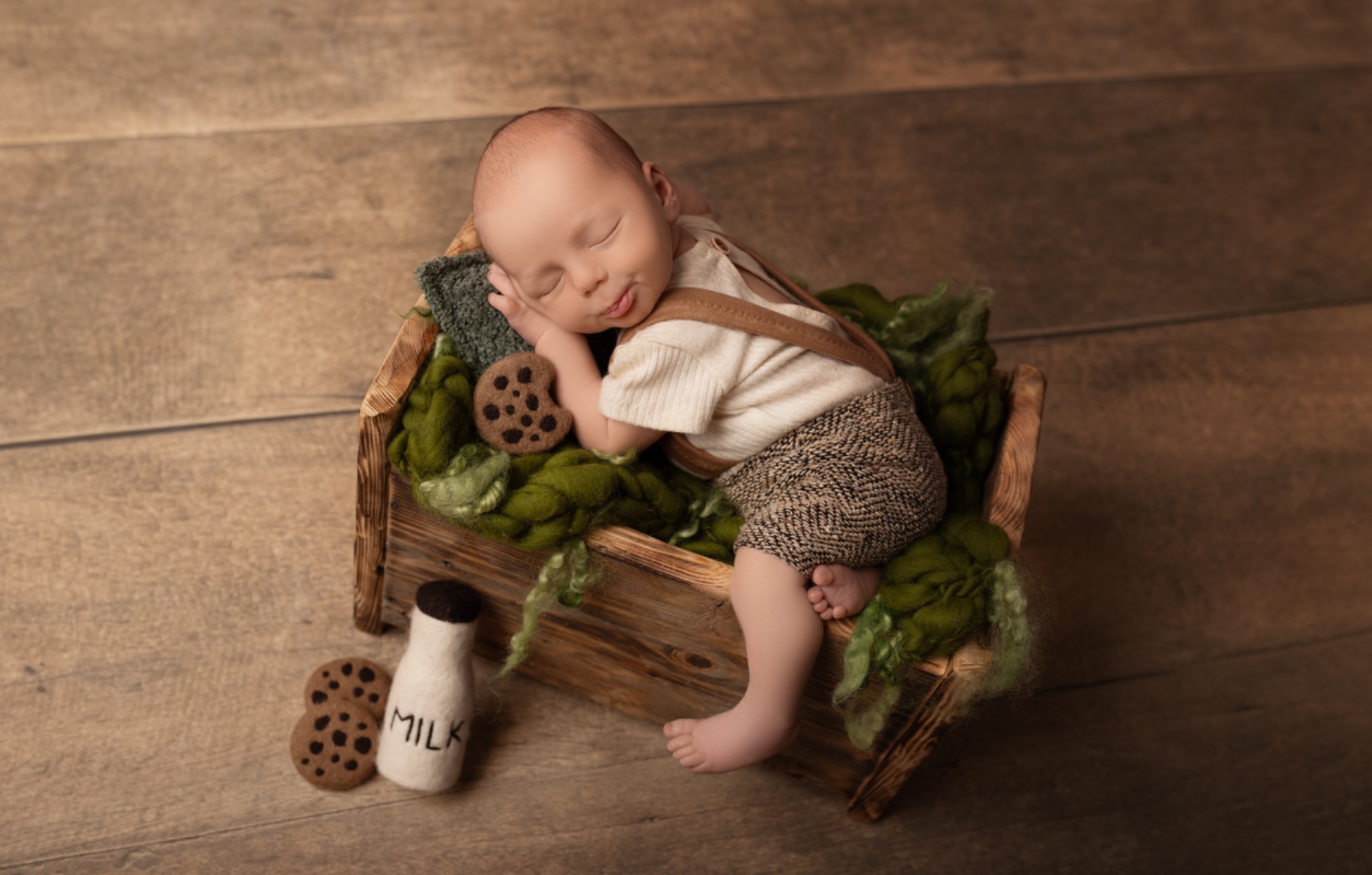 Noworodek śpiący w drewnianej skrzynce z zielonym wypełnieniem, obok butelka mleka i ciasteczka. Urocza stylizacja na sesję noworodkową.
