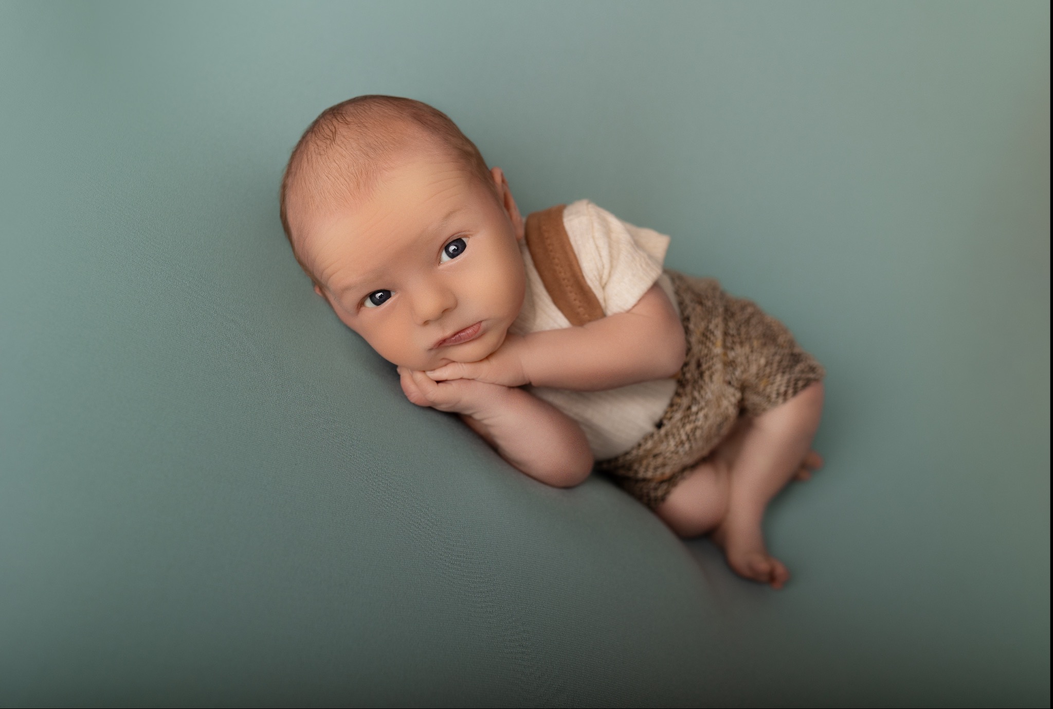 Noworodek w brązowym ubranku leży na boku na turkusowym tle, opierając głowę na dłoniach. Uwagę przykuwają duże, niebieskie oczy dziecka.