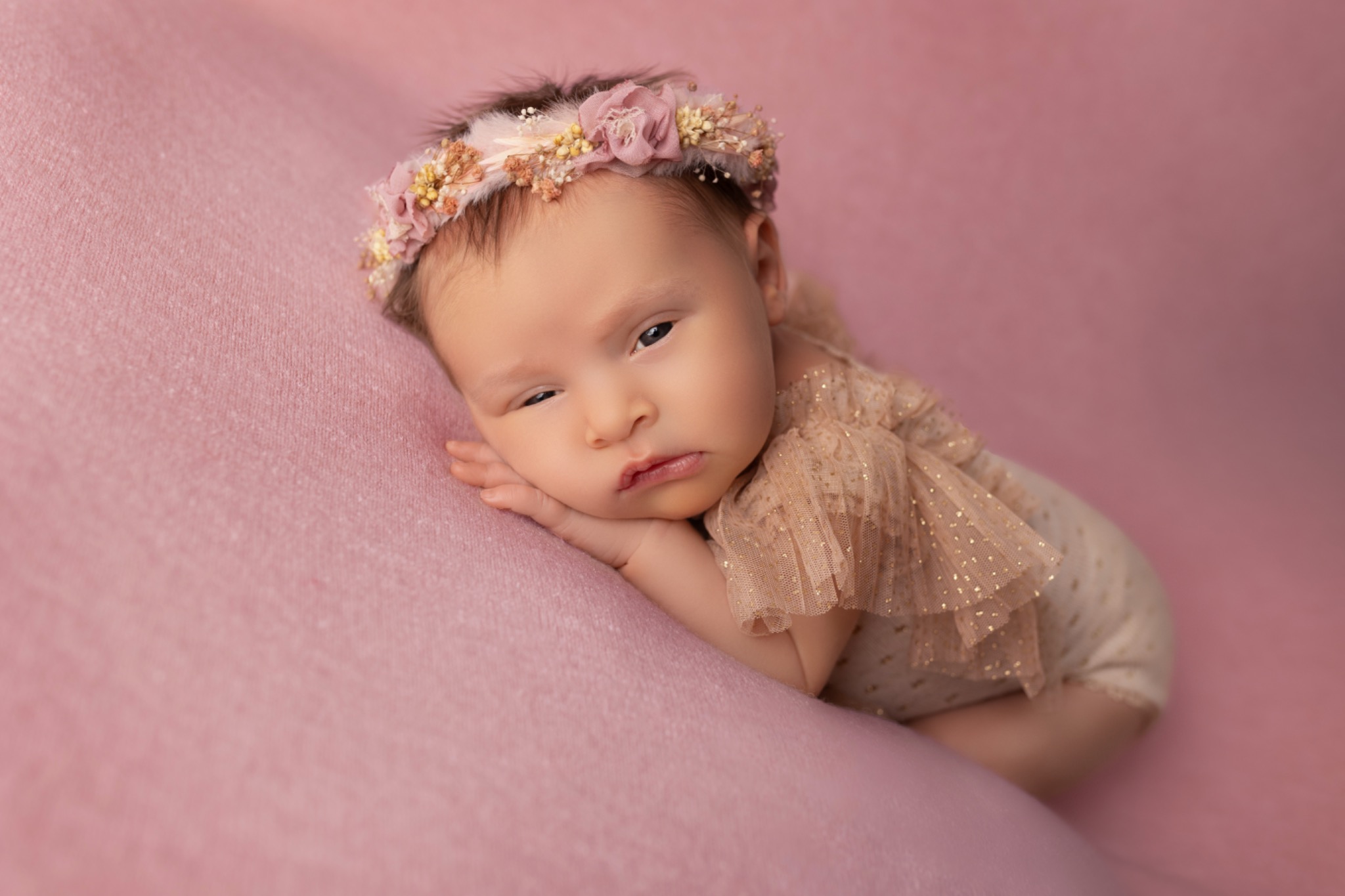 Noworodek w beżowym stroju z falbankami i kwiatowym wiankiem na głowie, leżący na różowym tle. Dziecko ma otwarte jedno oko i lekko zmarszczone usta.
