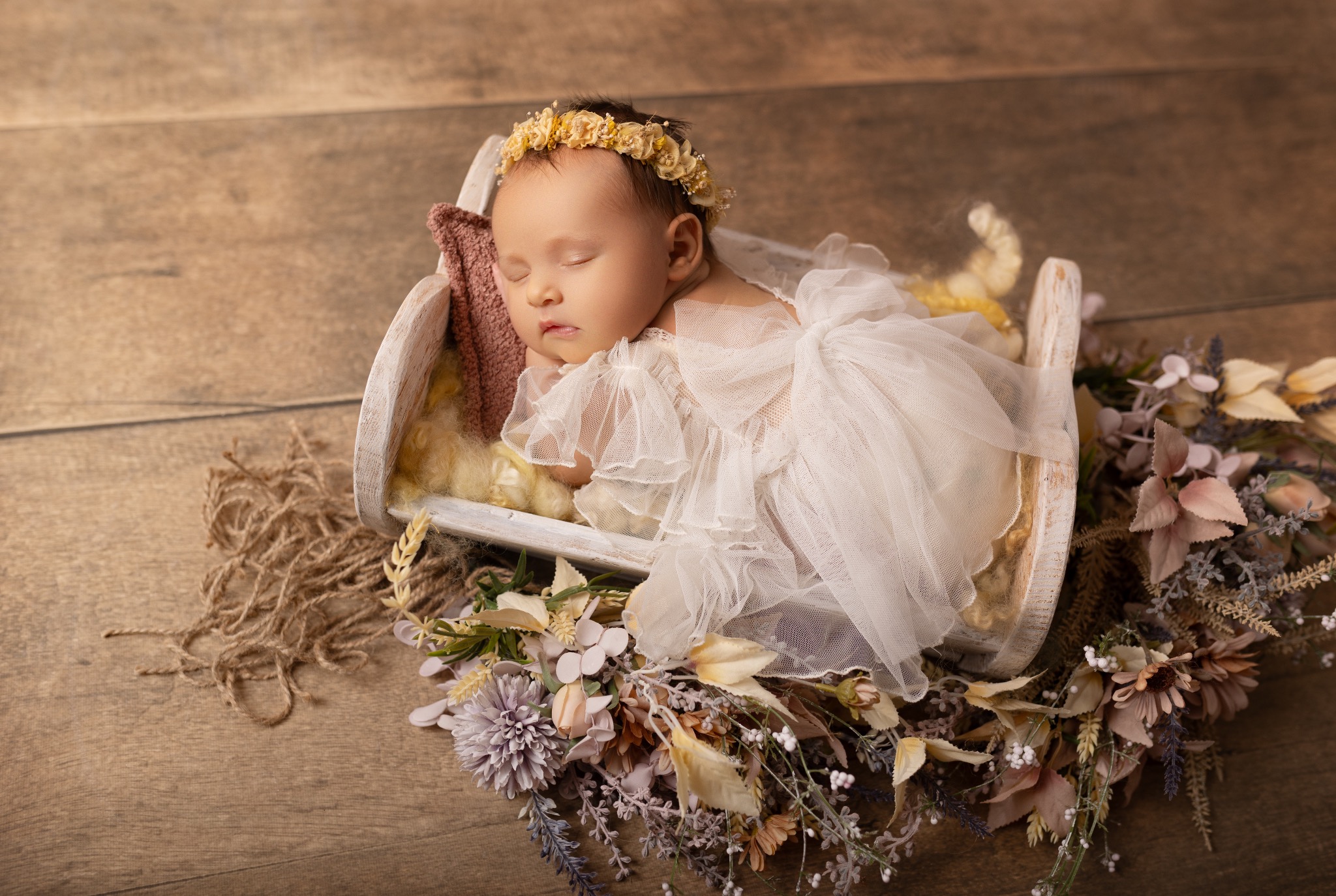 Śpiąca noworodka w białej sukience i wianku, ułożona w drewnianym łóżeczku z kwiatami na drewnianym tle. Delikatne, naturalne światło.
