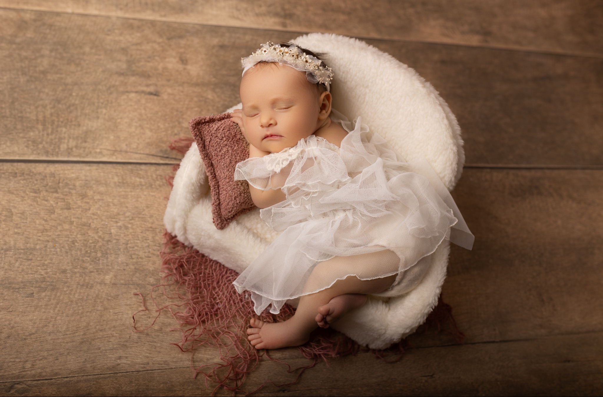 Śpiąca noworodka w białej sukience i opasce z kwiatami, ułożona w miękkim koszyku na drewnianym tle. Urocza sesja w ciepłych barwach.