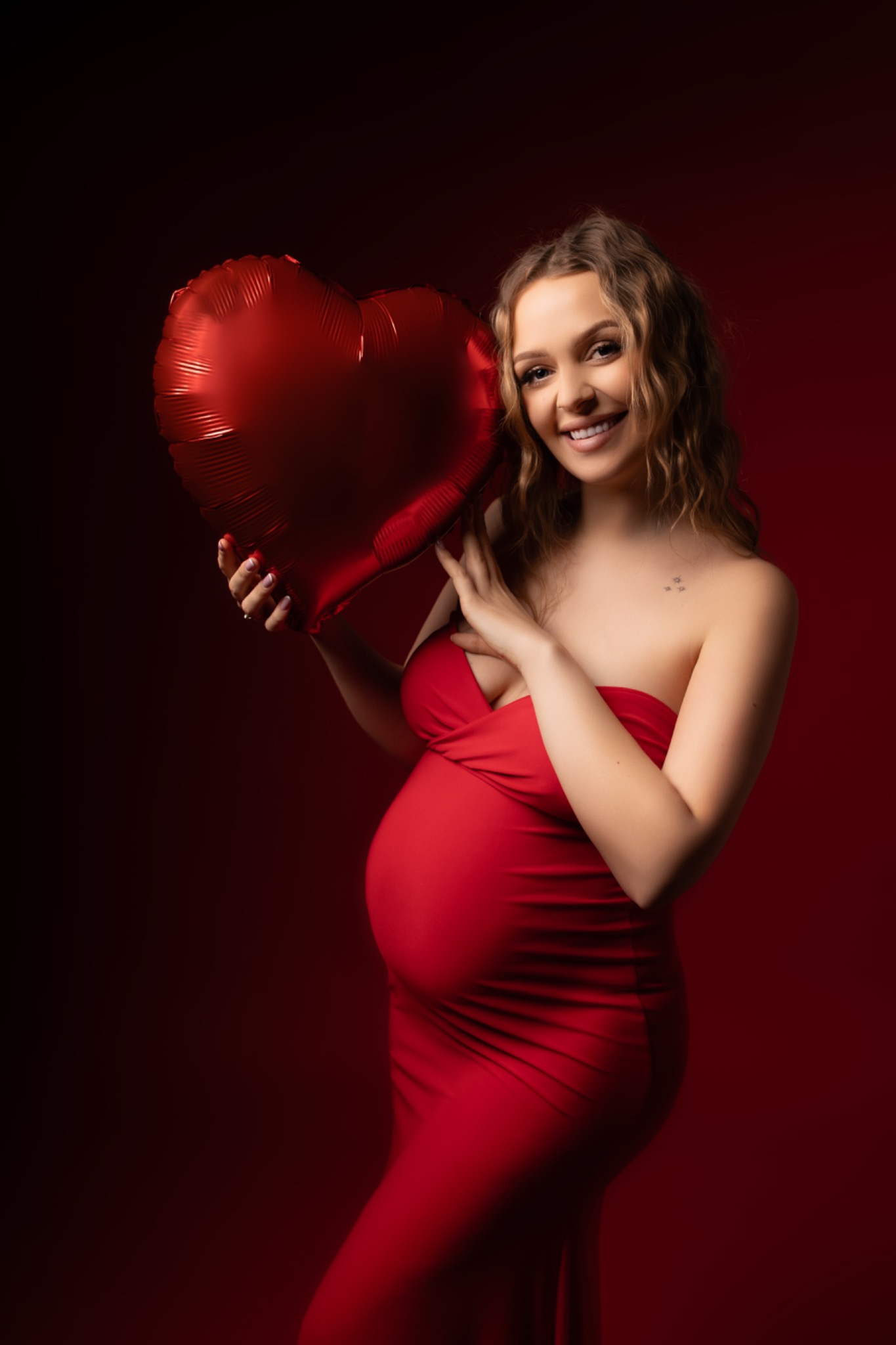 Uśmiechnięta, ciężarna blondynka w czerwonej sukni trzyma czerwony balon w kształcie serca na ciemnoczerwonym tle. Romantyczna sesja ciążowa.
