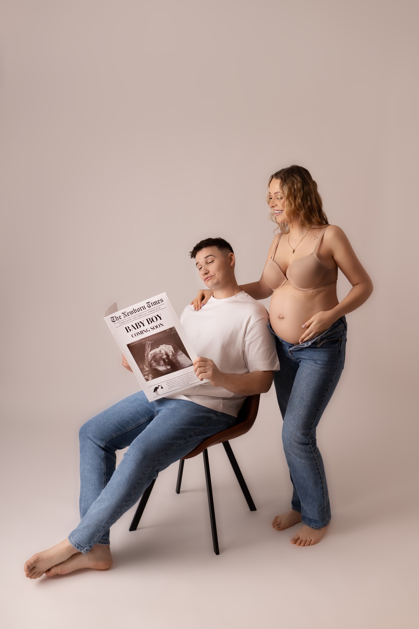 Uśmiechnięta, ciężarna kobieta w jeansach i biustonoszu opiera się na ramieniu mężczyzny czytającego gazetę 'Baby Boy Coming Soon' na jasnym tle.