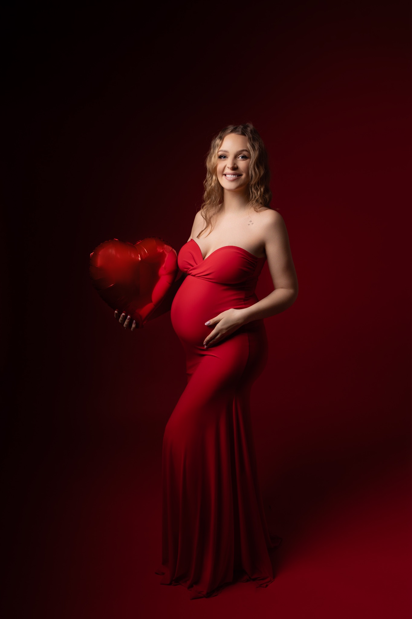 Uśmiechnięta kobieta w czerwonej sukni ciążowej, trzymająca balon w kształcie serca na ciemnoczerwonym tle. Widoczny brzuszek i tatuaż na ramieniu.