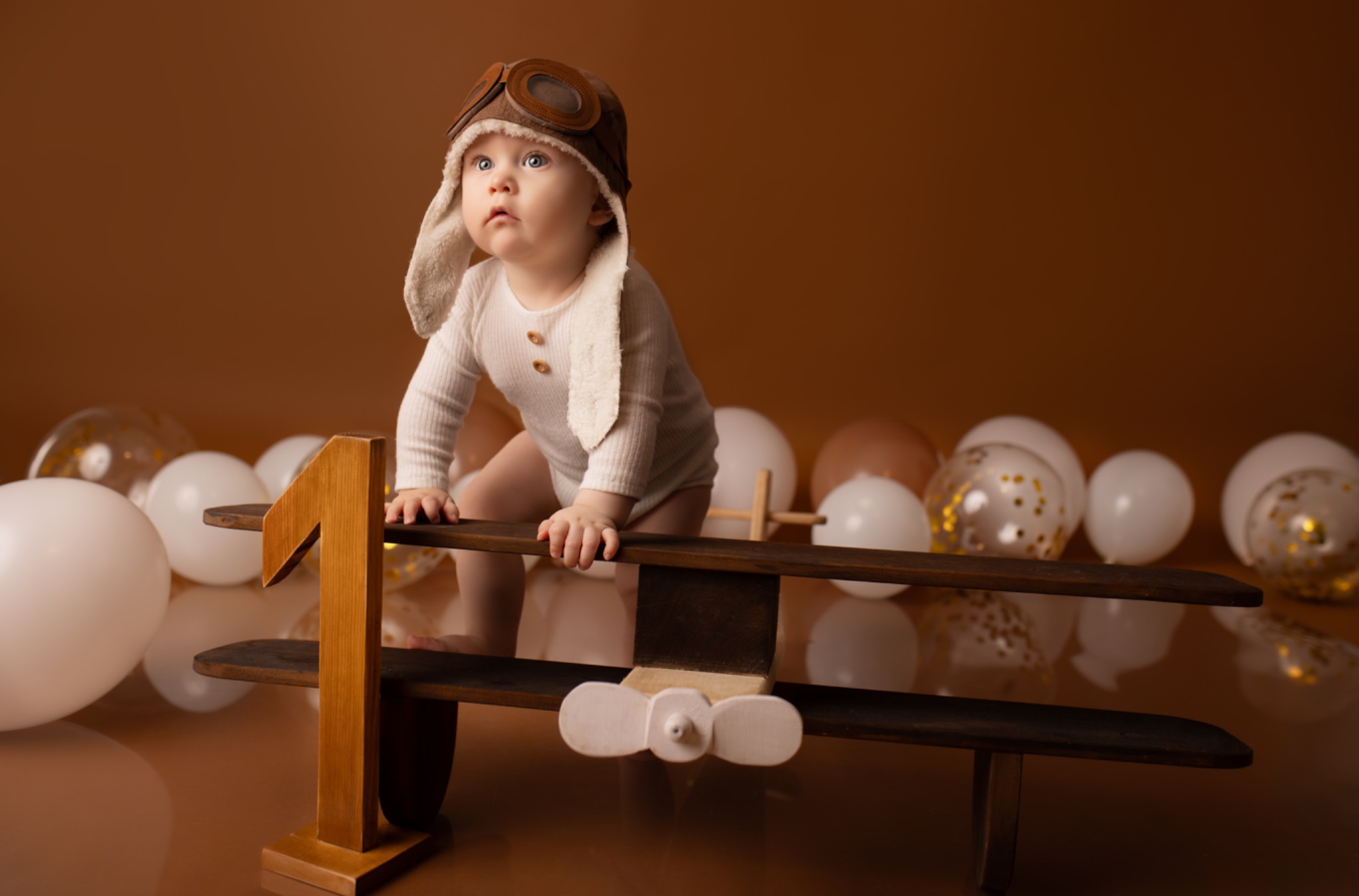 Urocze dziecko w czapce pilota pozuje na tle brązowego tła z drewnianym modelem samolotu i białymi balonami. Roczny chłopiec z zaciekawieniem spogląda w górę.